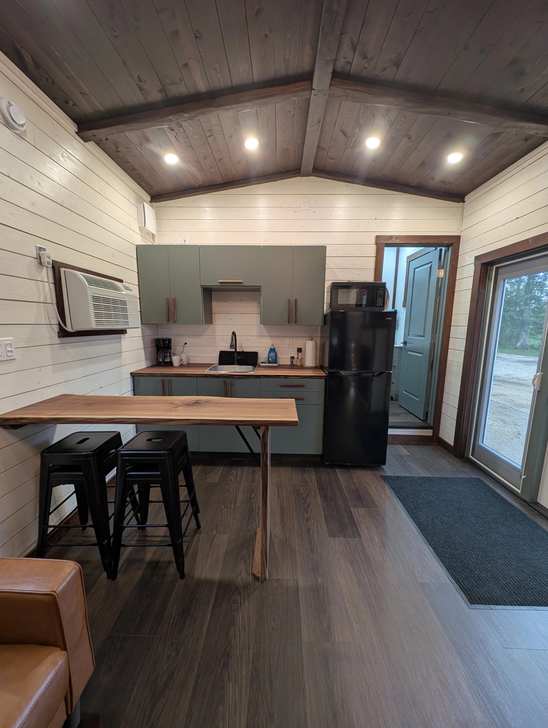 Compact kitchen with green cabinets, black appliances, wooden counter and stools.