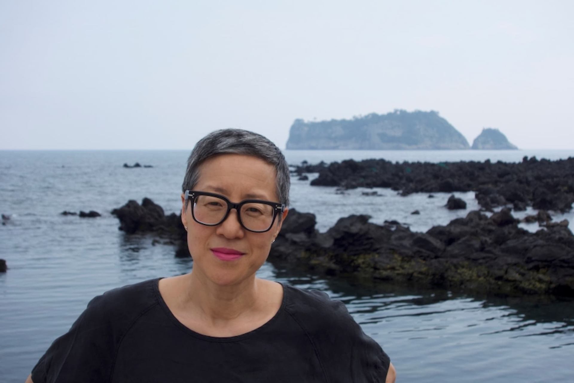 Person with short hair and glasses stands by rocky shoreline with distant island.