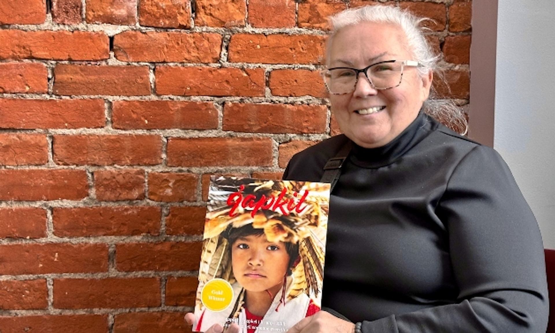 Person holding a colorful magazine with bold red title against a backdrop of exposed brick wall.