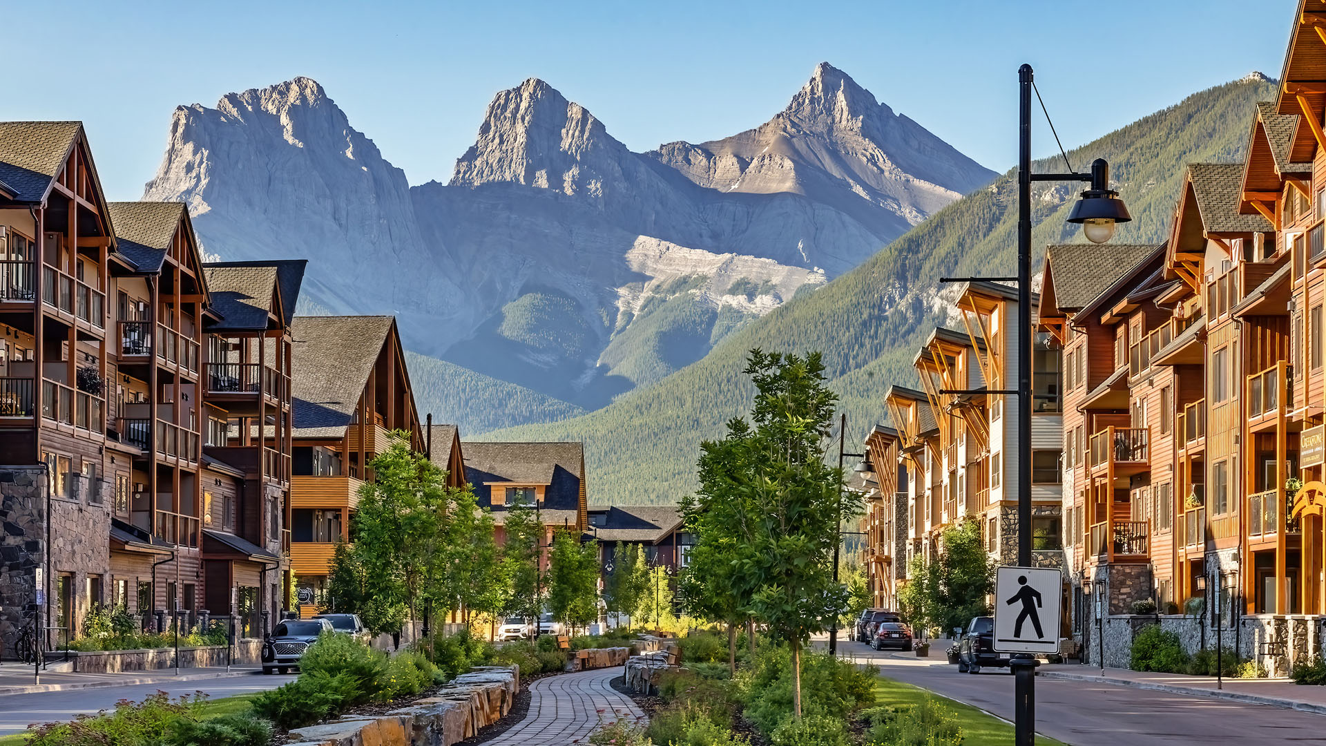 Mountain town street with wooden condos, trees, parked cars and towering rocky peaks.