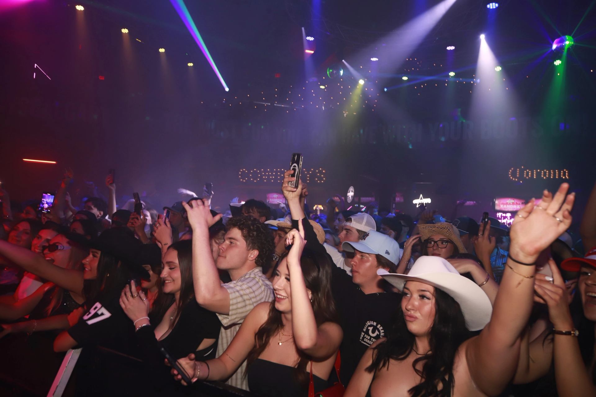 Crowd at indoor concert raises phones and hands under colourful stage lights.