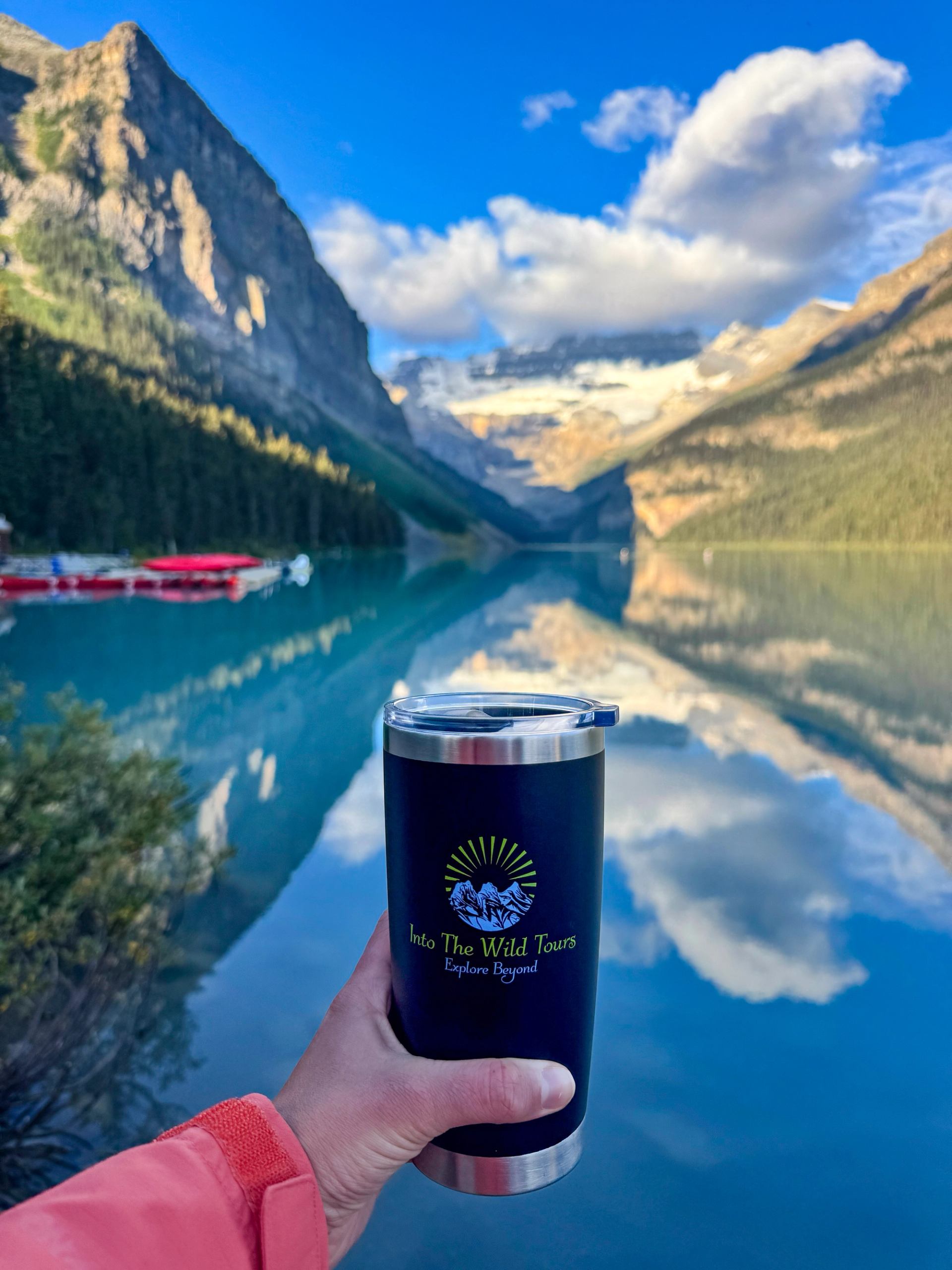 Hand holding an Into the Wild mug in front of Lake Louise’s calm waters.