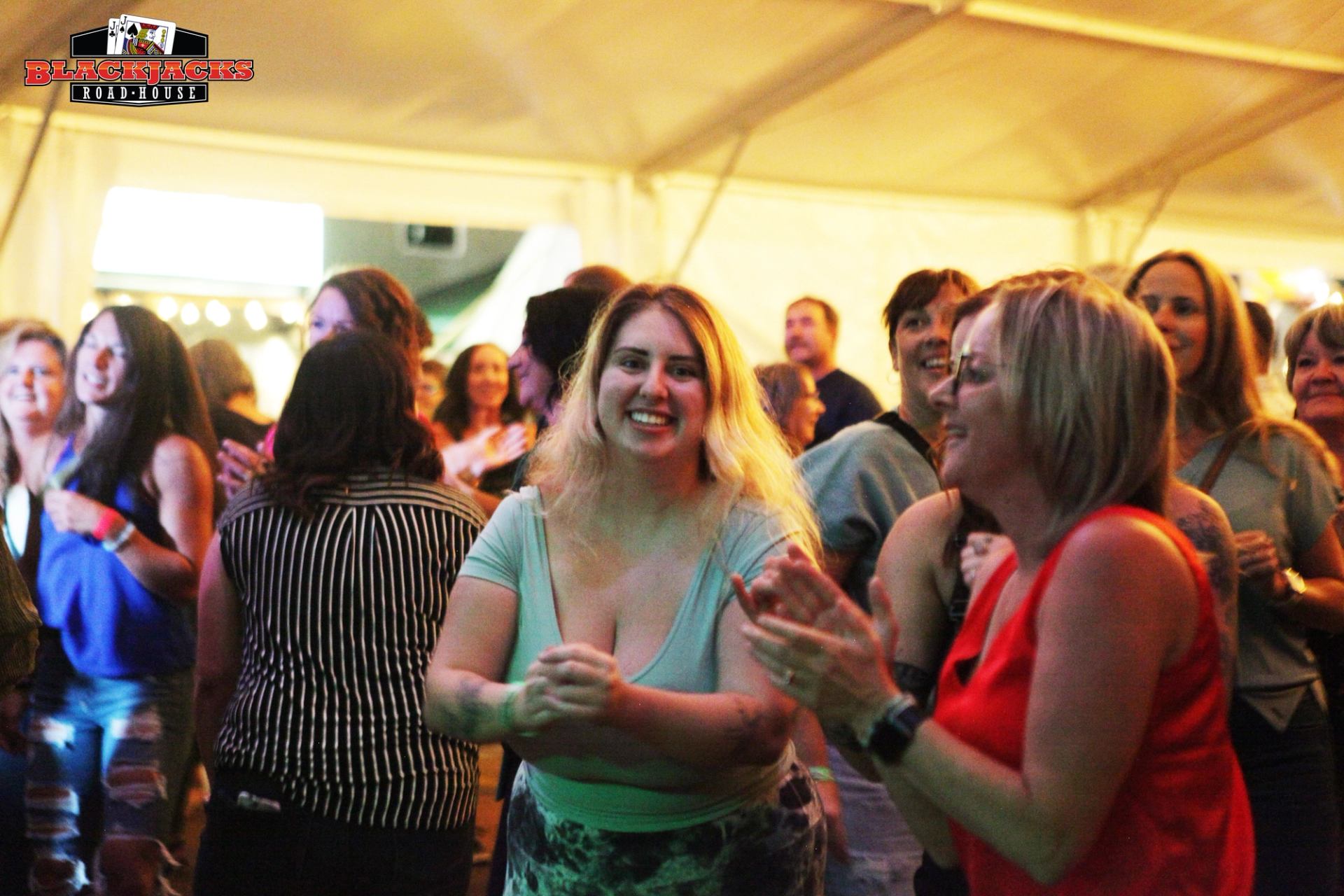 Crowd at a live music concert under a white tent, clapping and holding drinks.
