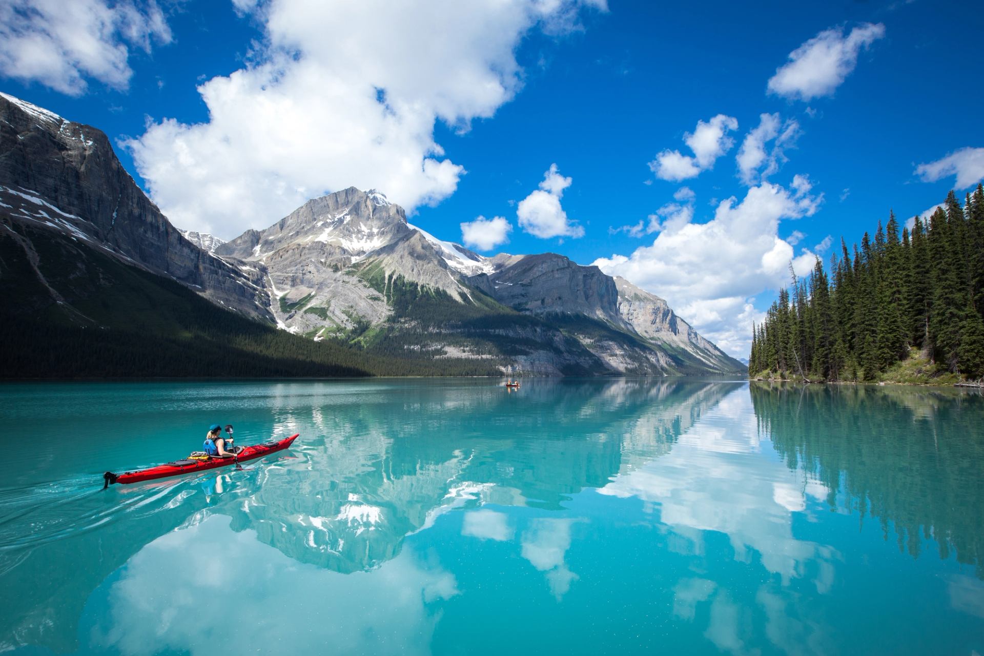 Red kayak glides across turquoise mountain lake, reflecting peaks and scattered clouds.
