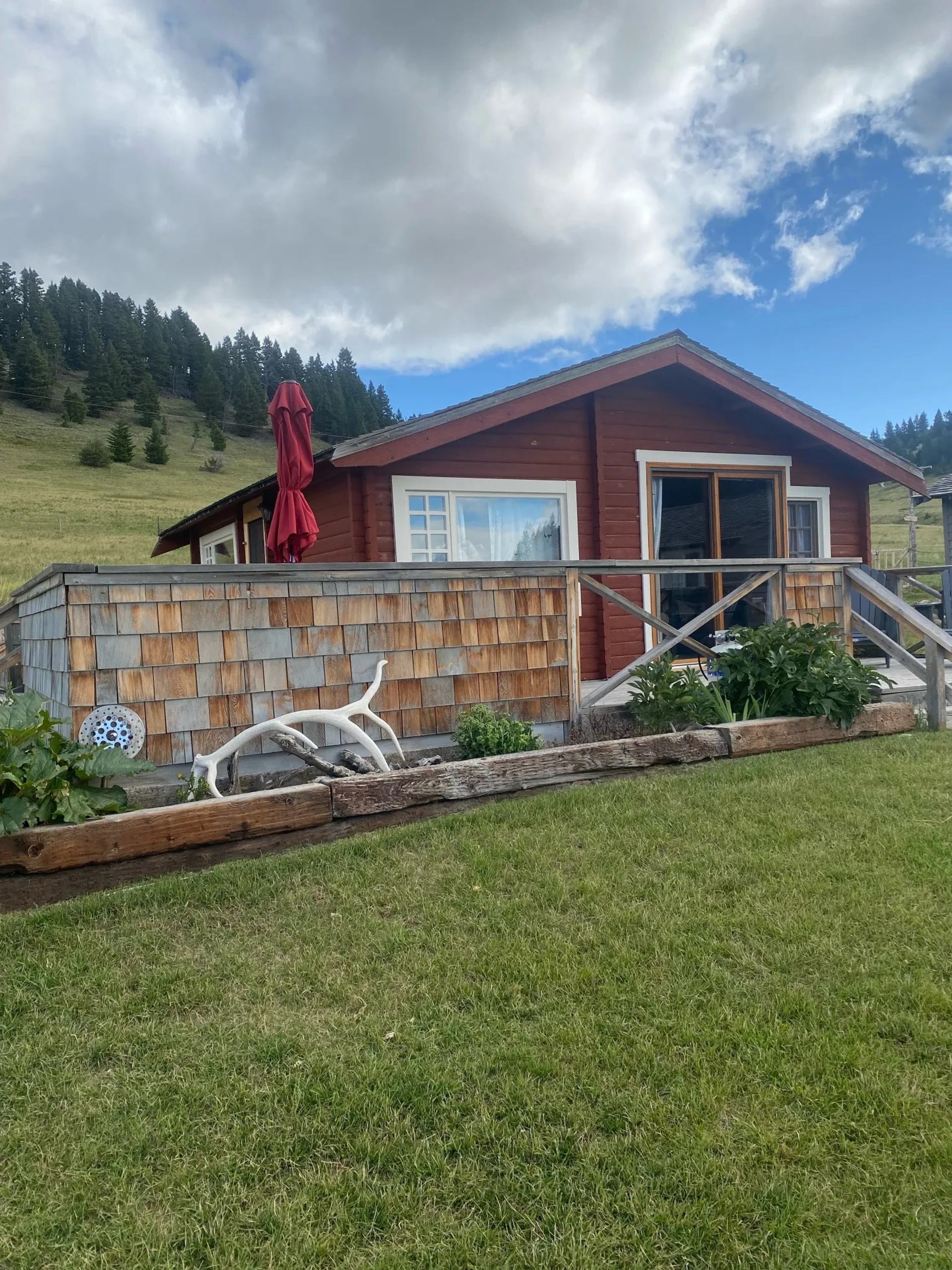 Red ranch cabin with shingle siding, wooden railing, and garden on a grassy hill under blue sky.