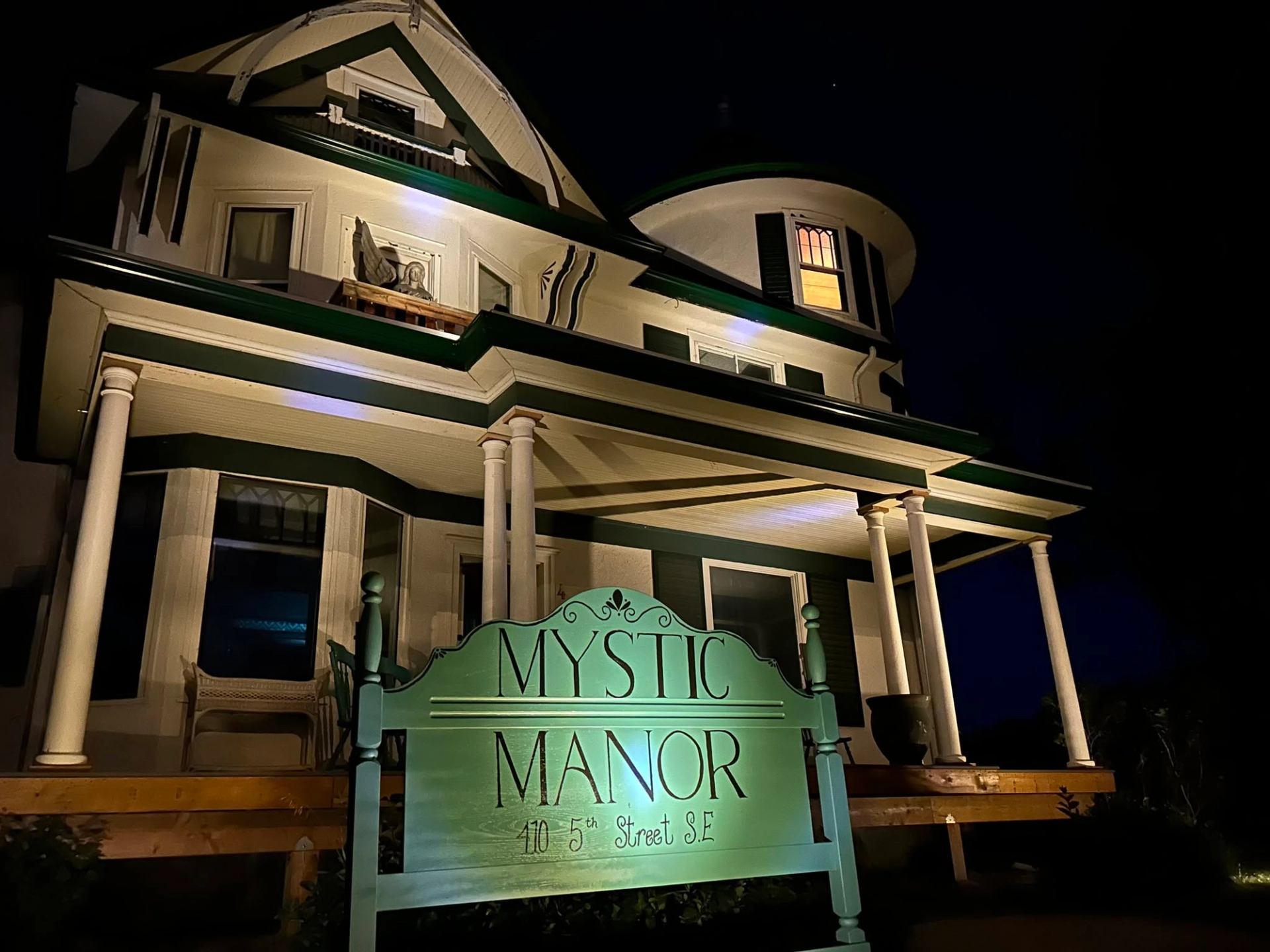 Historic house at night with lit Mystic Manor sign and wraparound porch.