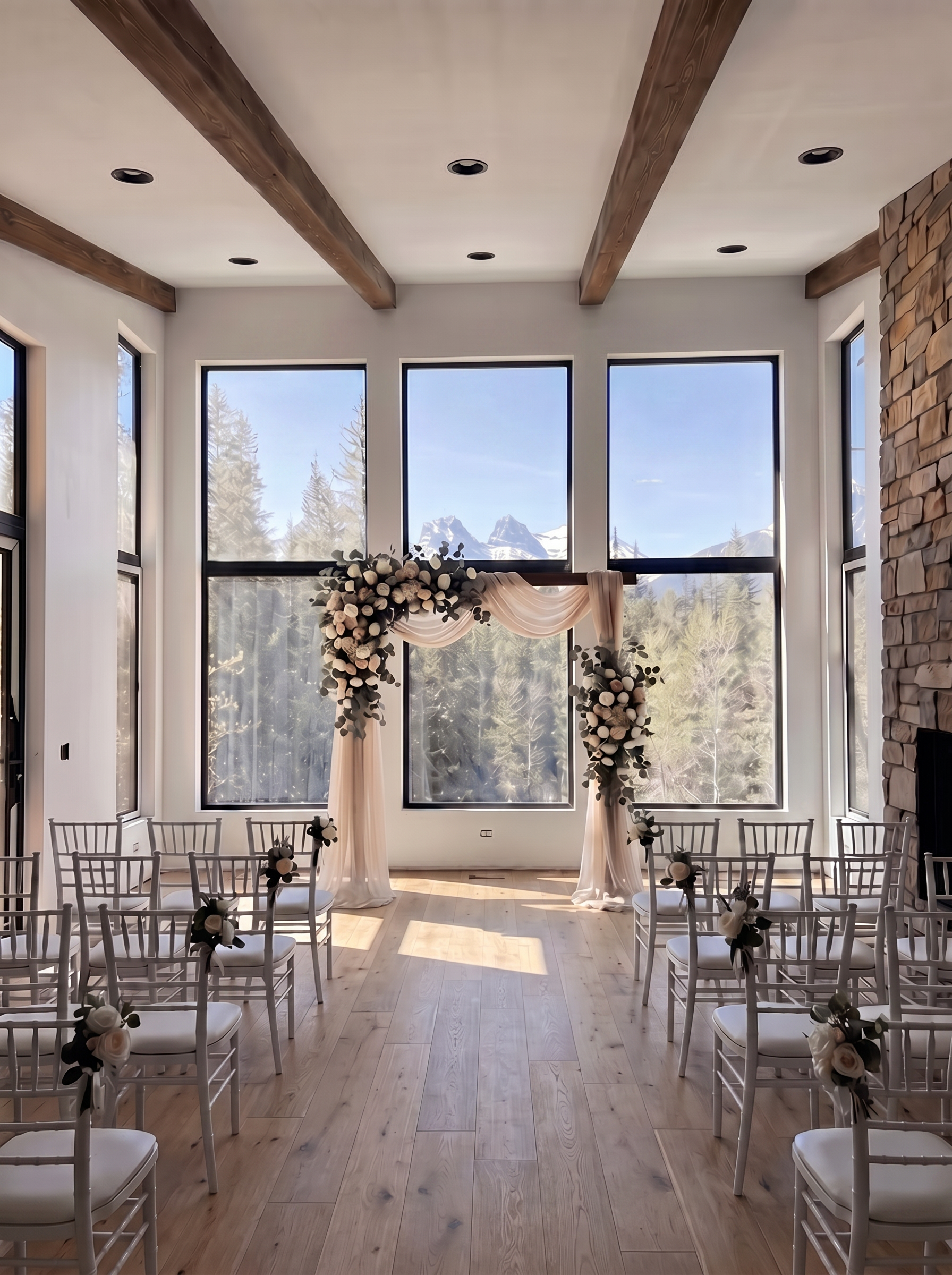 Indoor wedding ceremony setup with floral arch, white chairs and mountain view through windows.