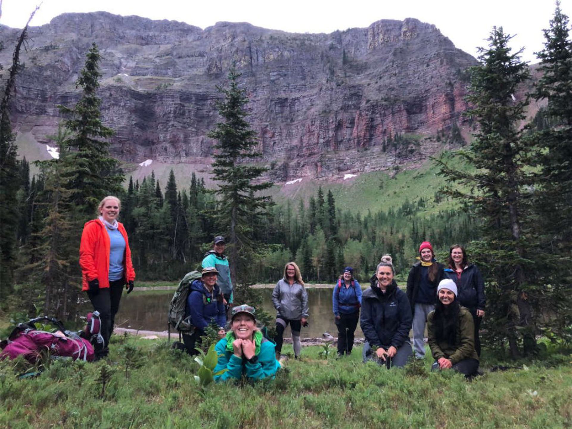 Backpackers gathered near a lake with steep mountain cliffs and trees in the background.