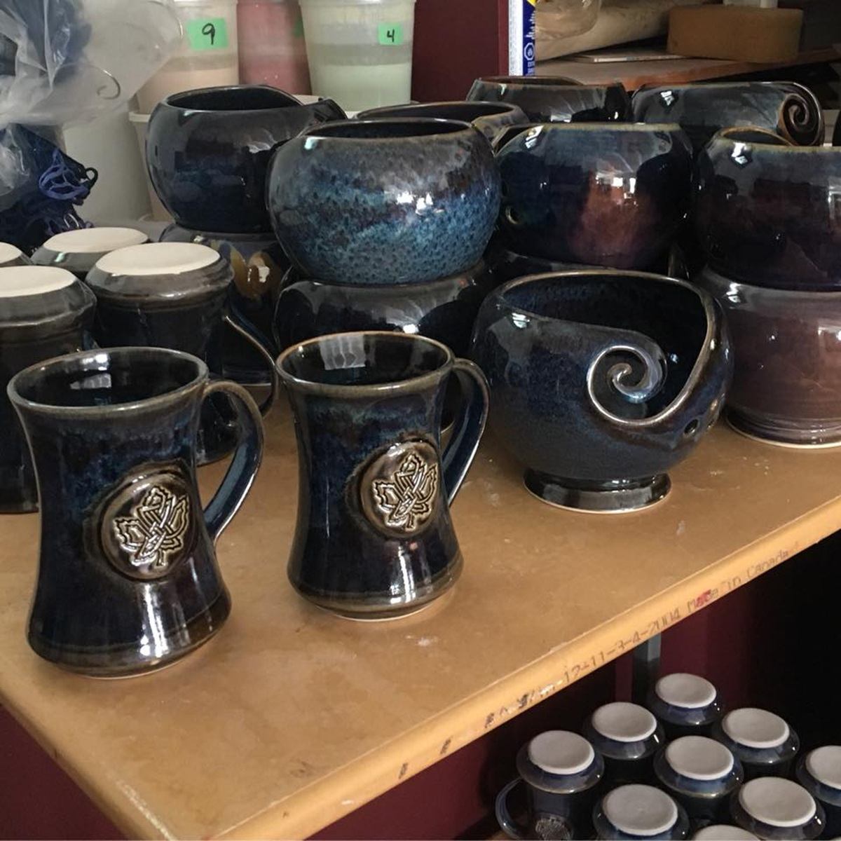 Dark blue handmade pottery mugs and bowls with maple leaf emblems on wooden shelves.