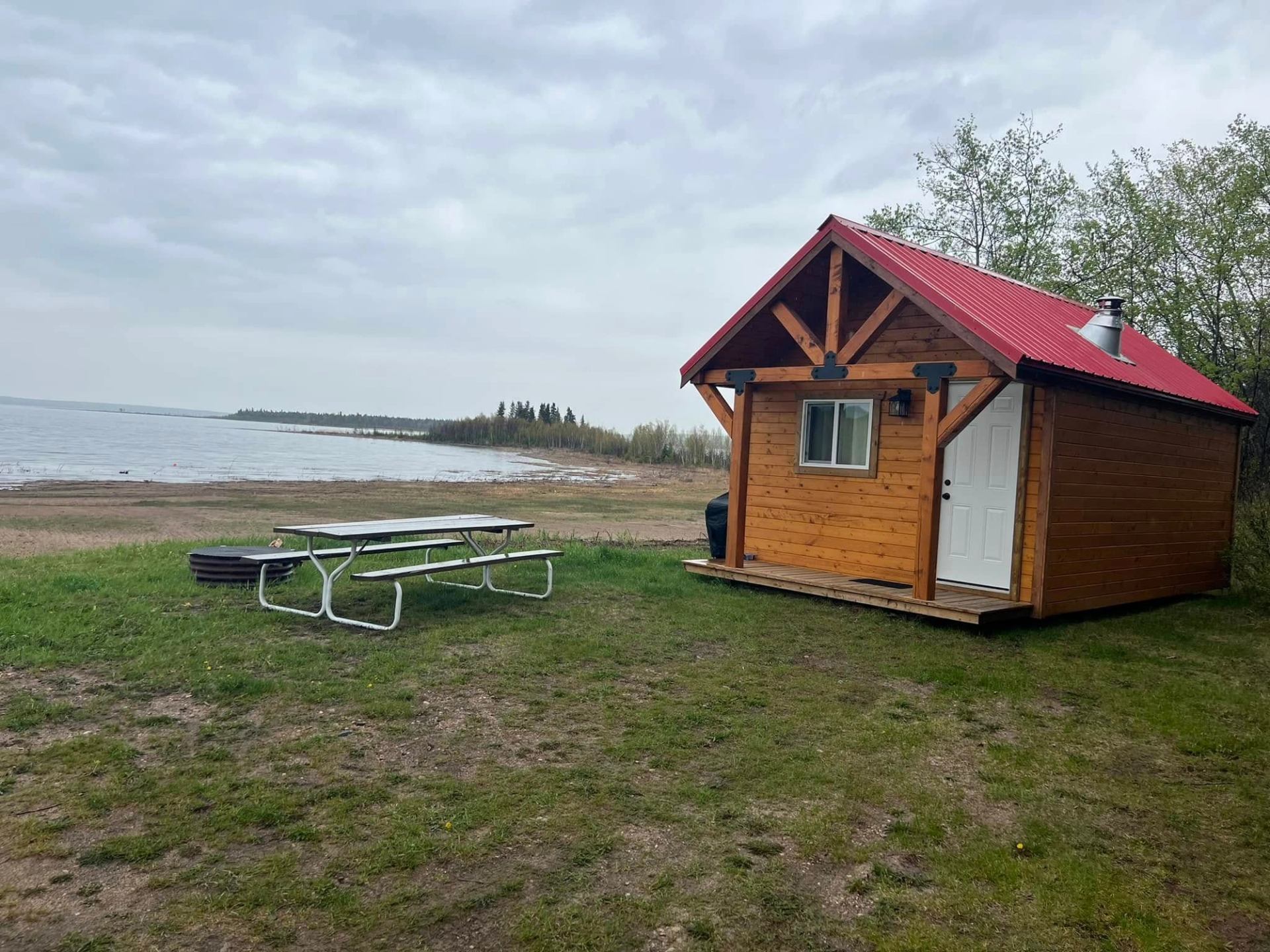 Small wooden cabin with red roof beside lake, picnic table and fire ring.