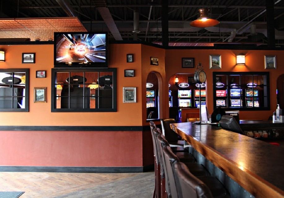 Warm bar interior with stools, framed art, TV, and slot machines in the back.