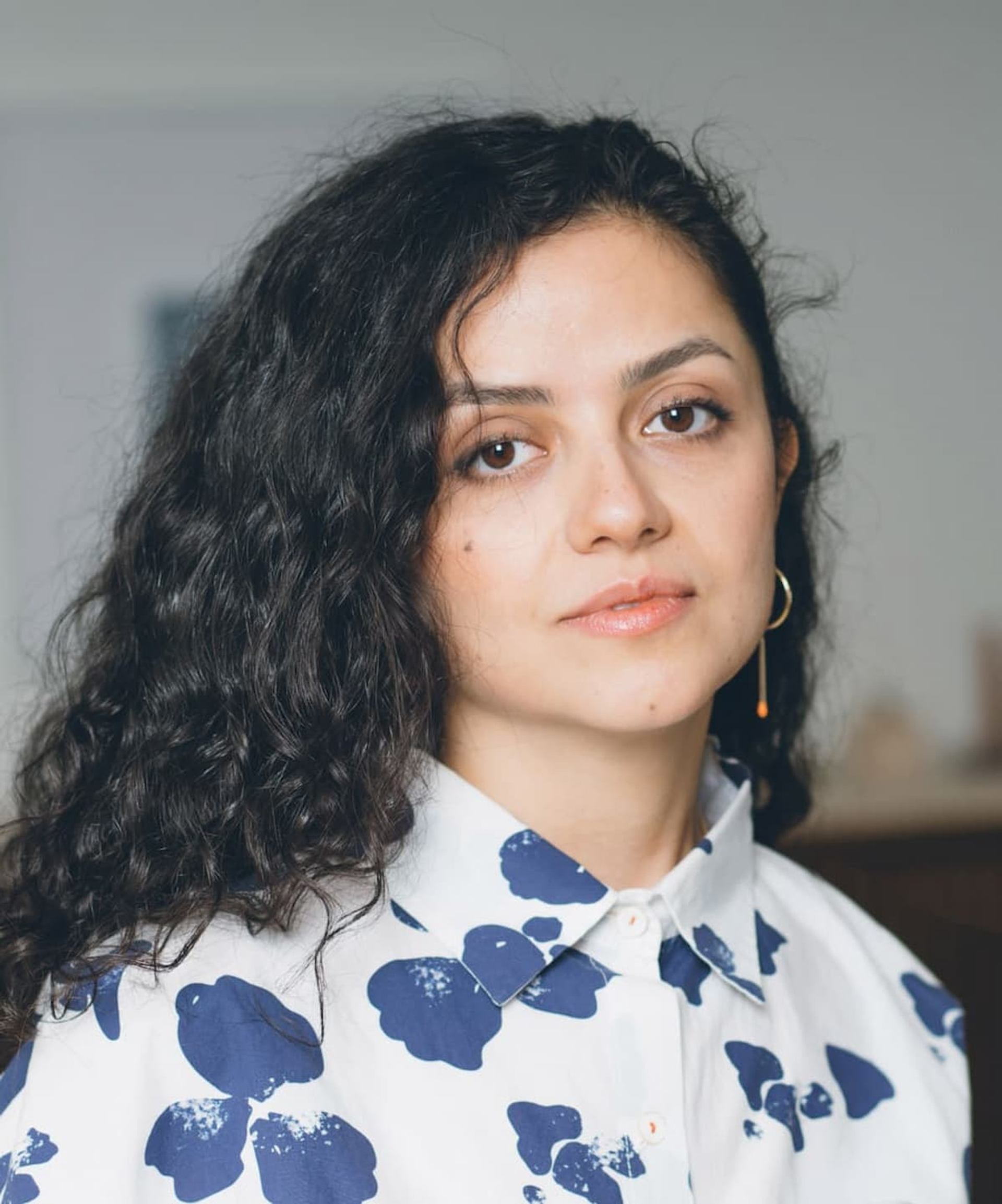 A woman with dark curly hair wears a white shirt with blue floral patterns and a gold drop earring.
