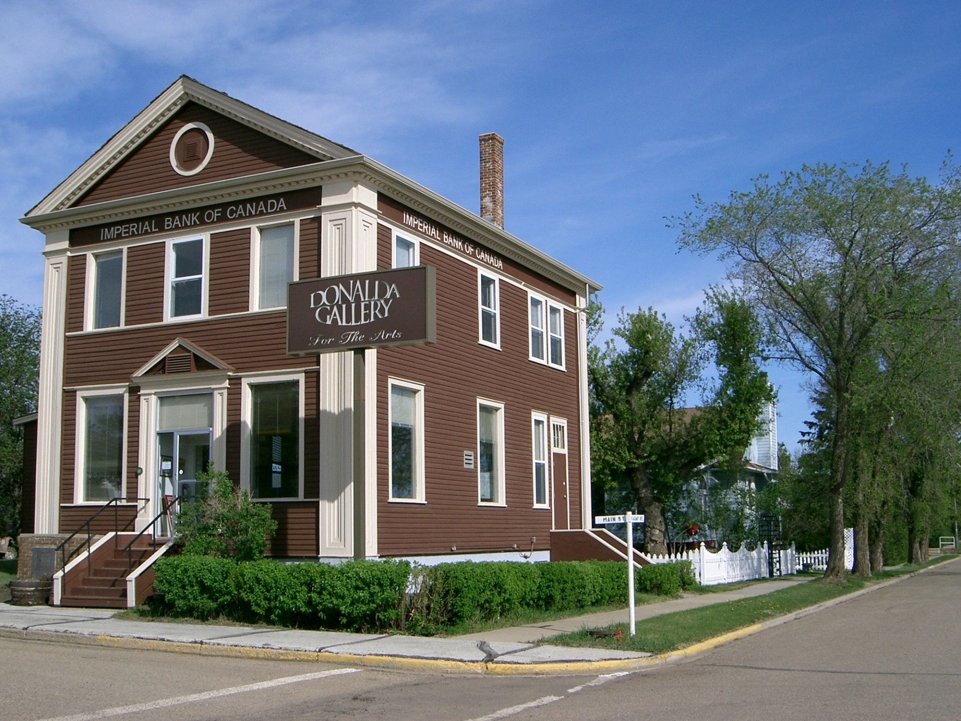 A heritage building with a sign reading "Donalda Gallery For The Arts" located on a street corner. The building has large windows, a staircase, and is surrounded by trees and green landscaping.