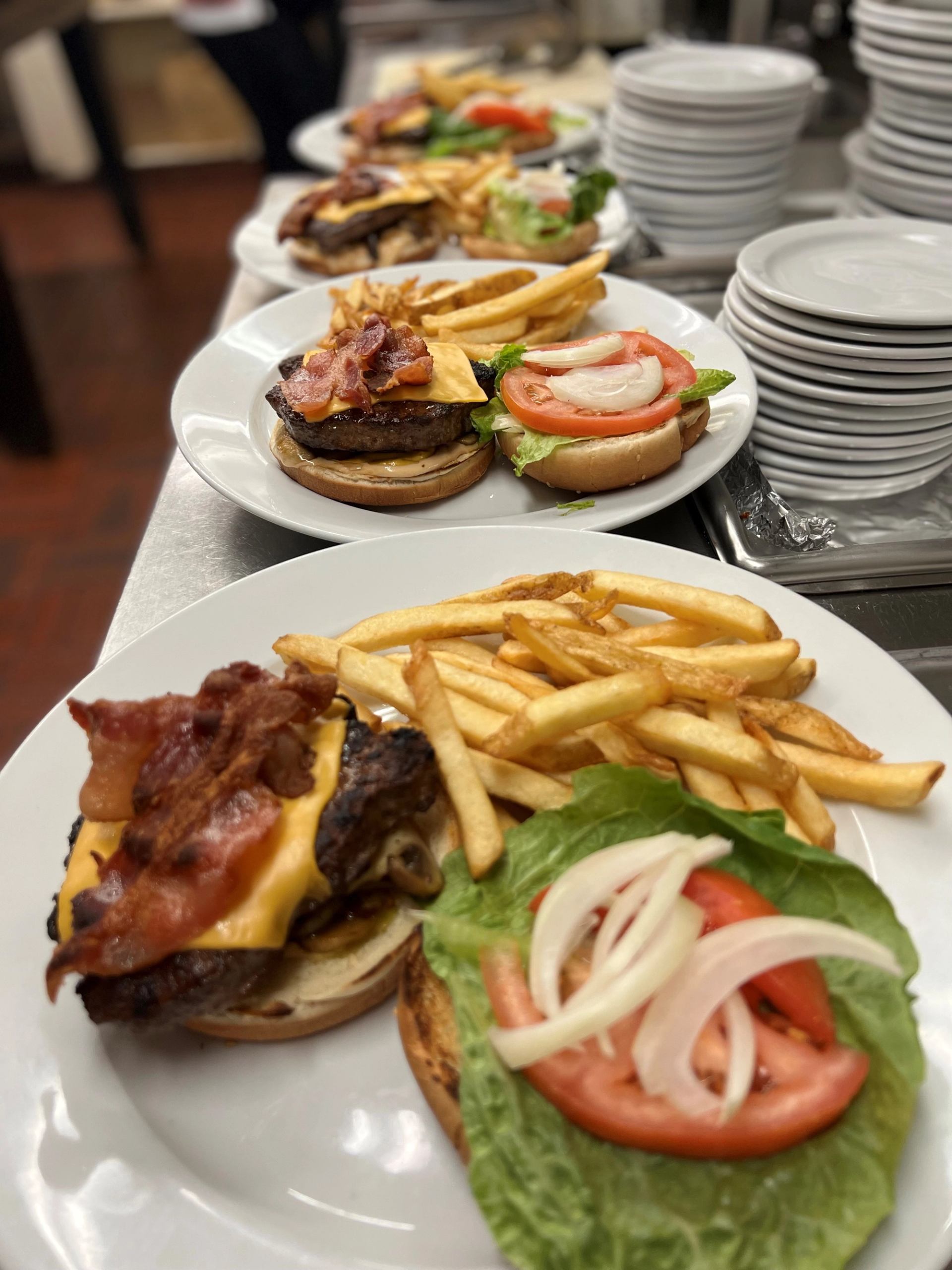 Plates of burgers and fries on a restaurant table with stacked white dishes in background.