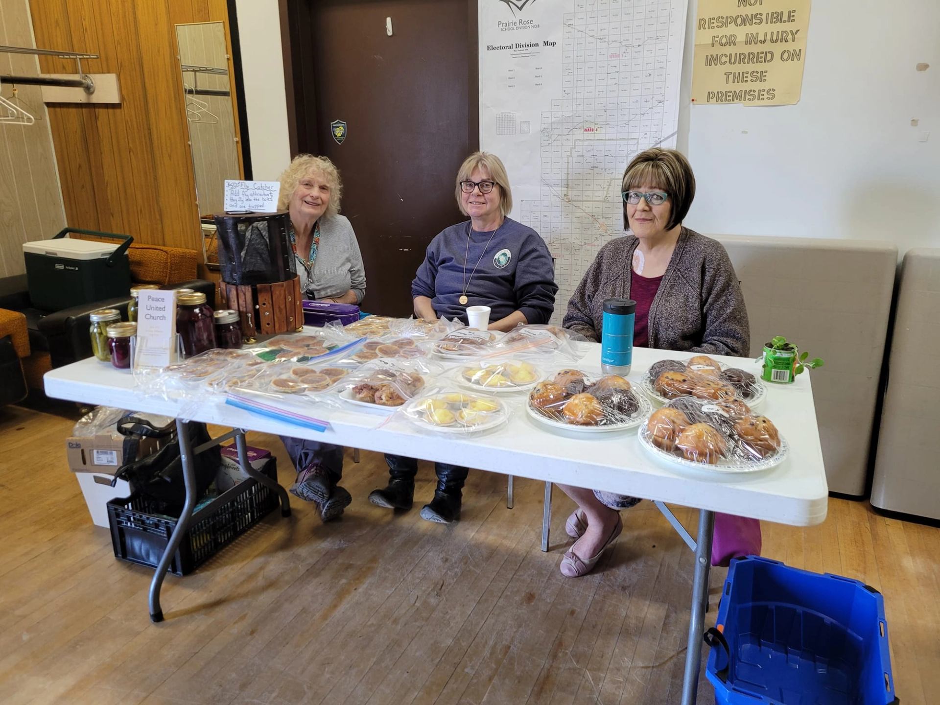 Three smiling women sit behind a table covered with baked goods and preserves.