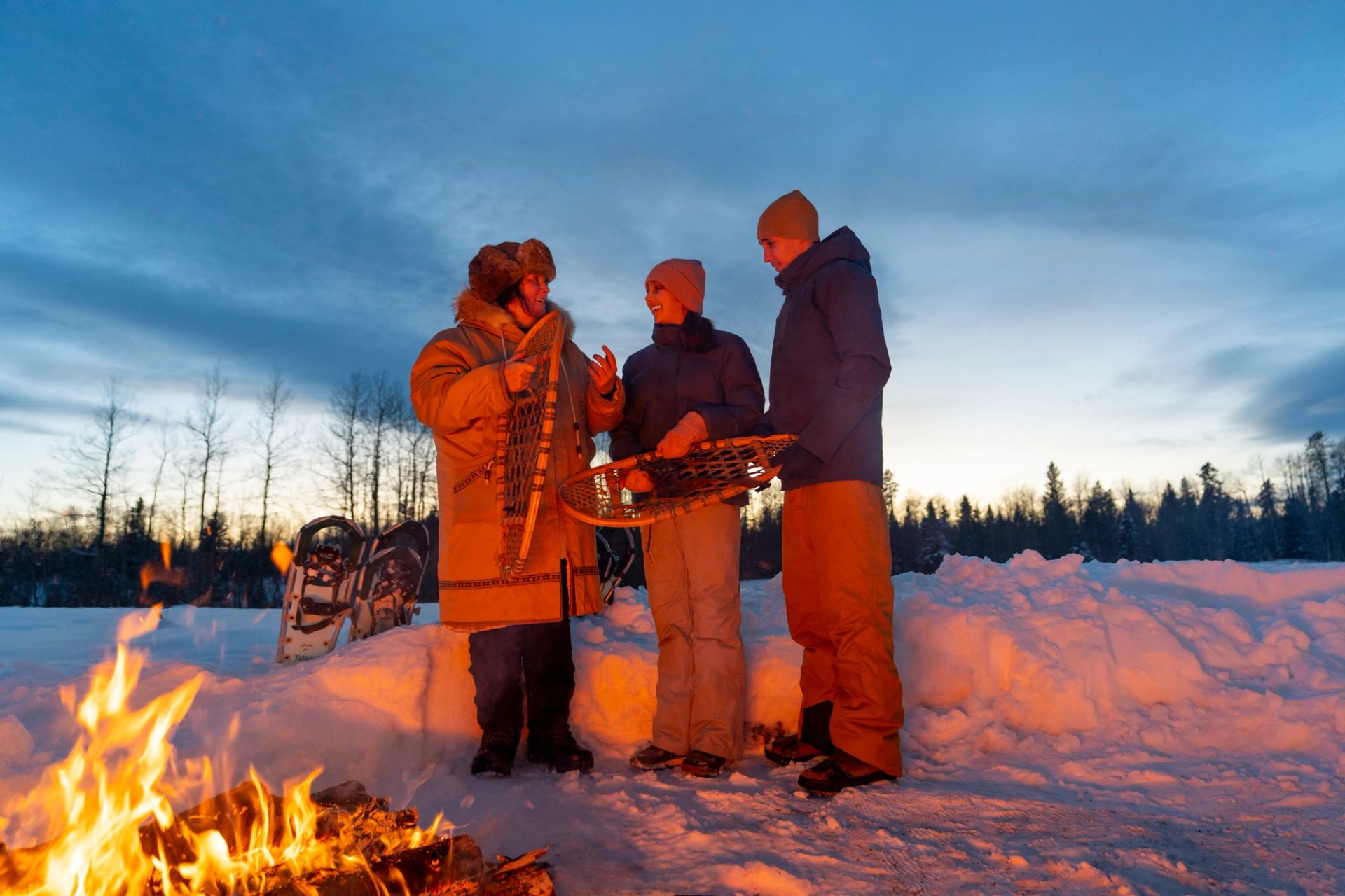 Three people standing near a campfire in the snow with snowshoes at sunset