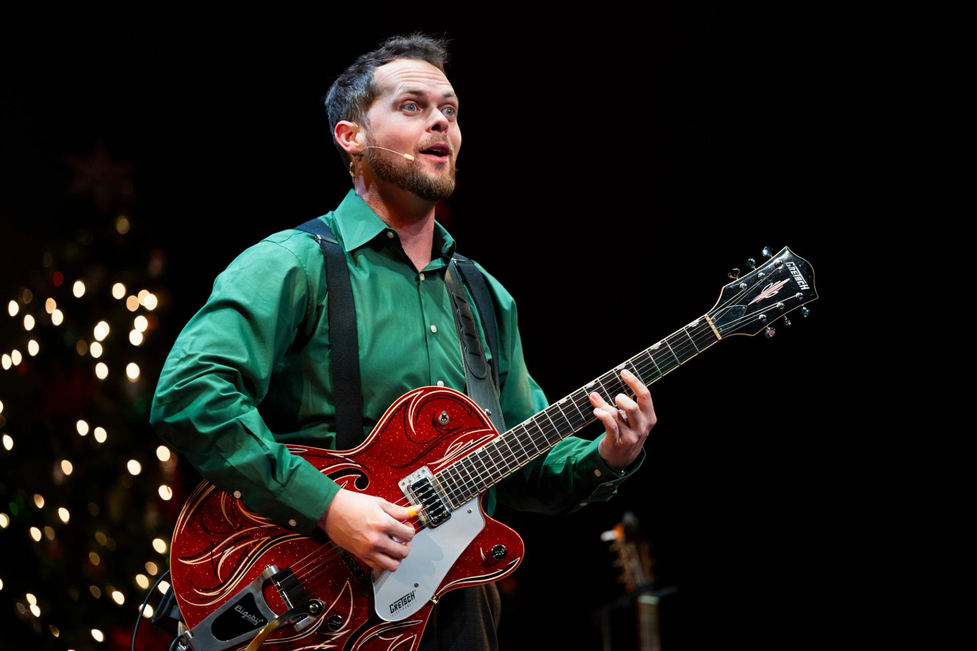 Guitarist in a green shirt playing a red electric guitar on stage with festive lights.