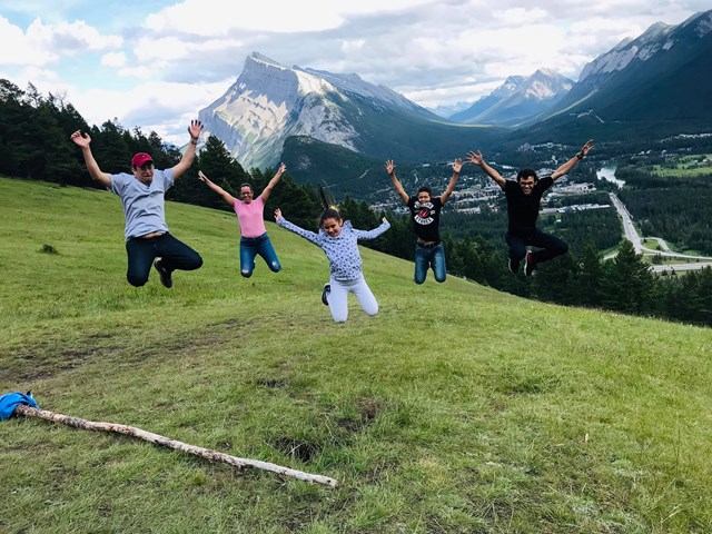 People jumping on grassy hillside, mountain peaks and valley visible in background.