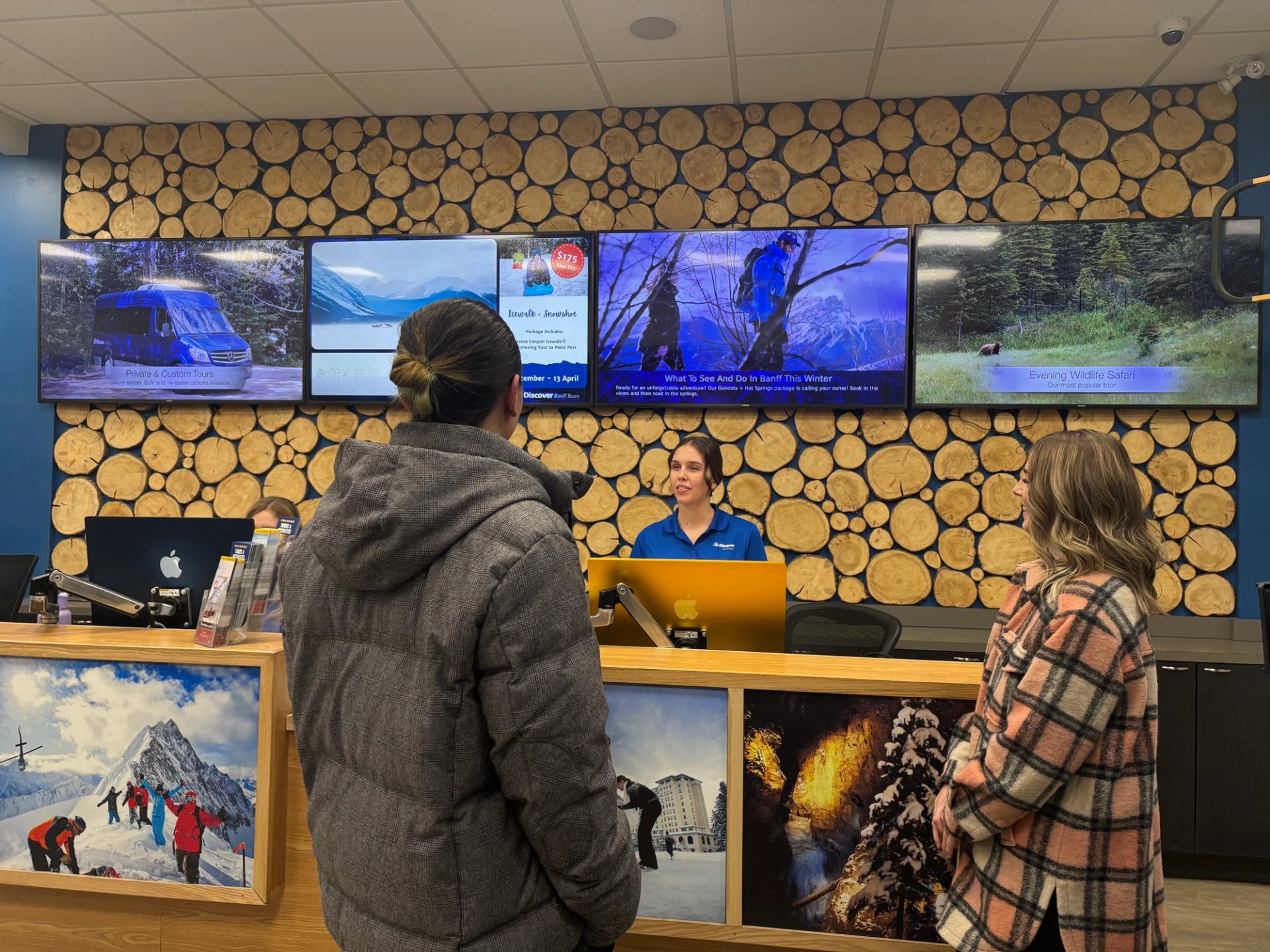 Guests speaking with staff at Discover Banff Tours desk with scenic images and screens.