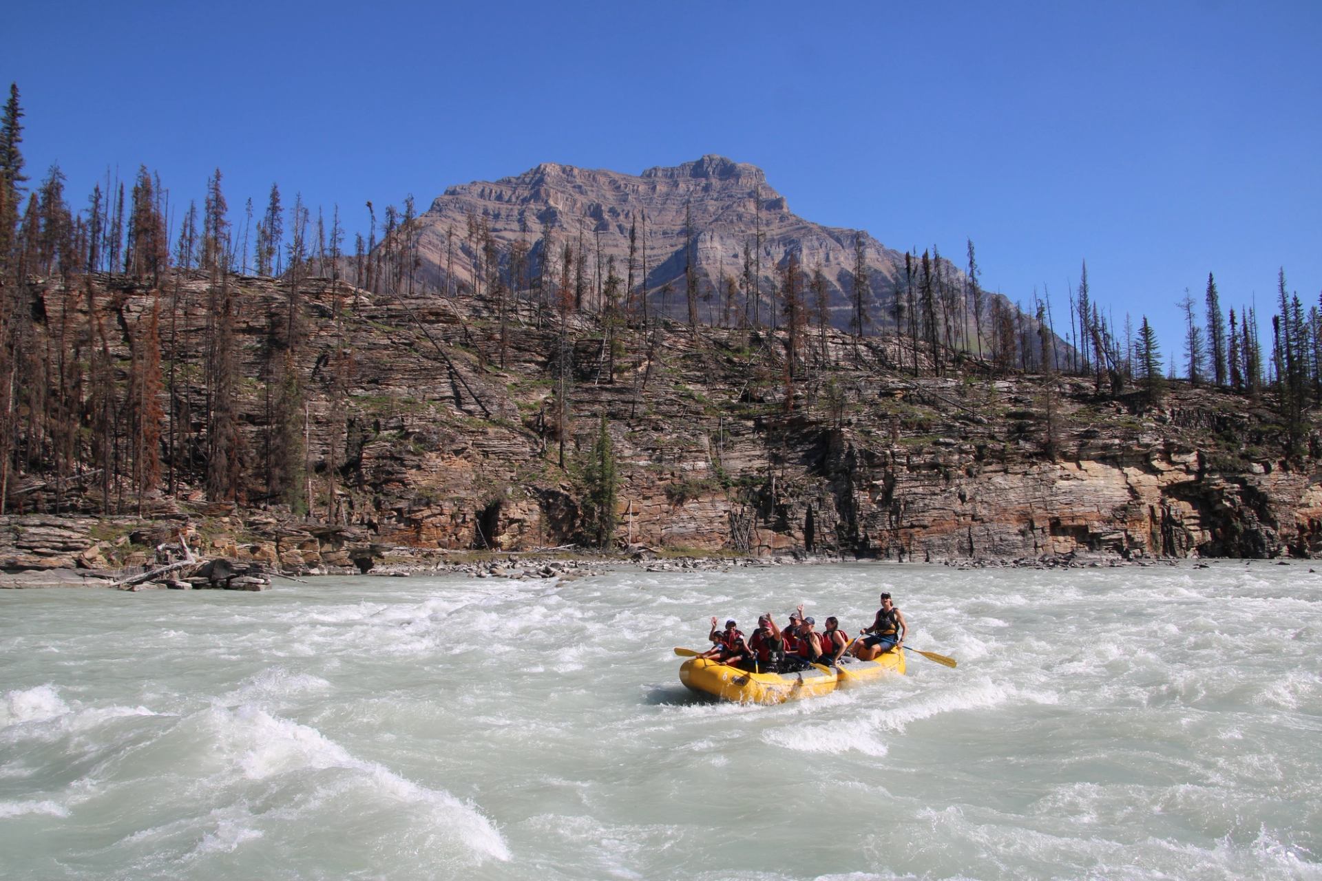 Yellow raft navigating swift whitewater with rocky cliffs and a large mountain in the background.