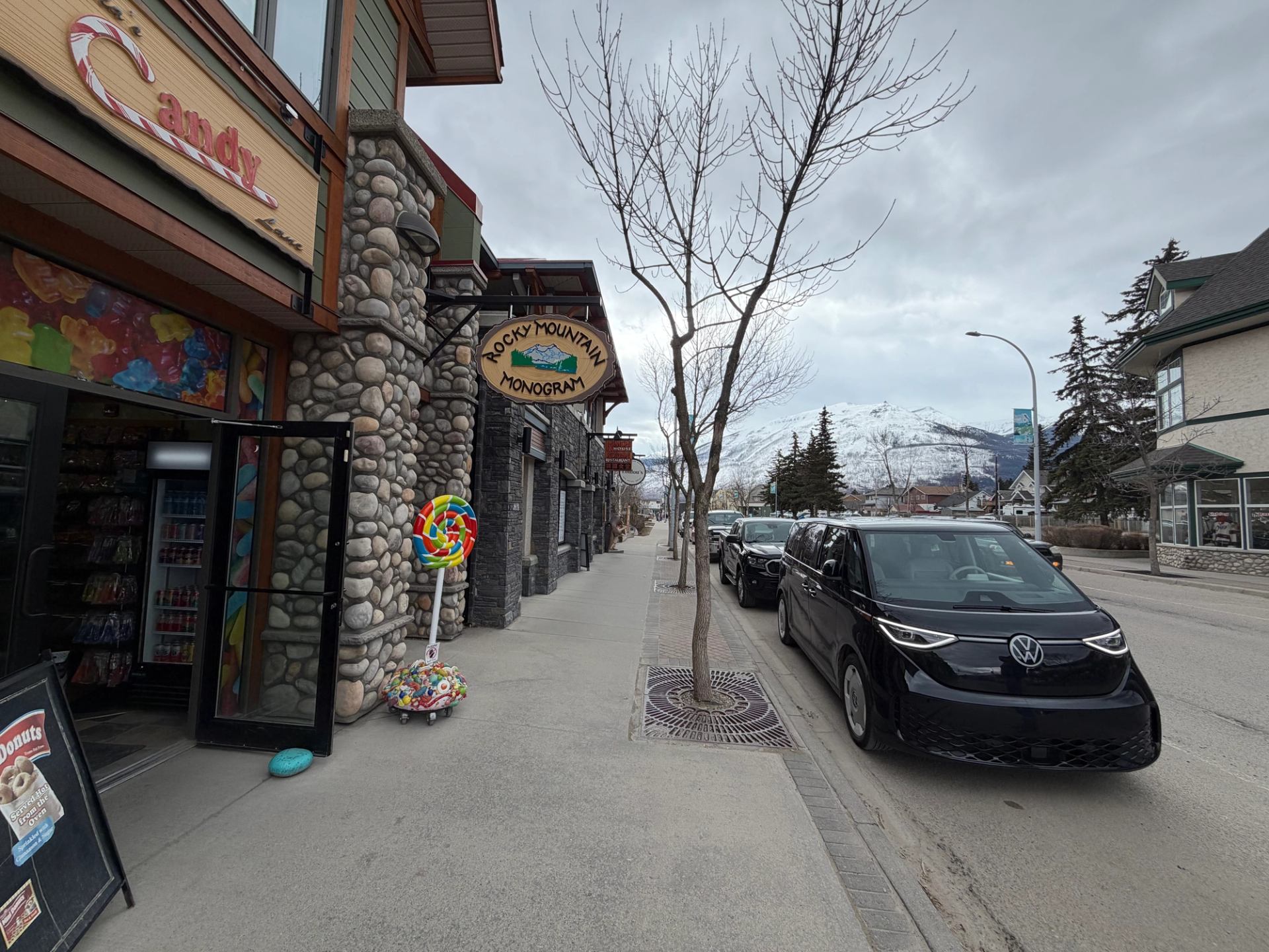 Street with candy shop, parked Volkswagen van, bare trees and snowy mountain backdrop.