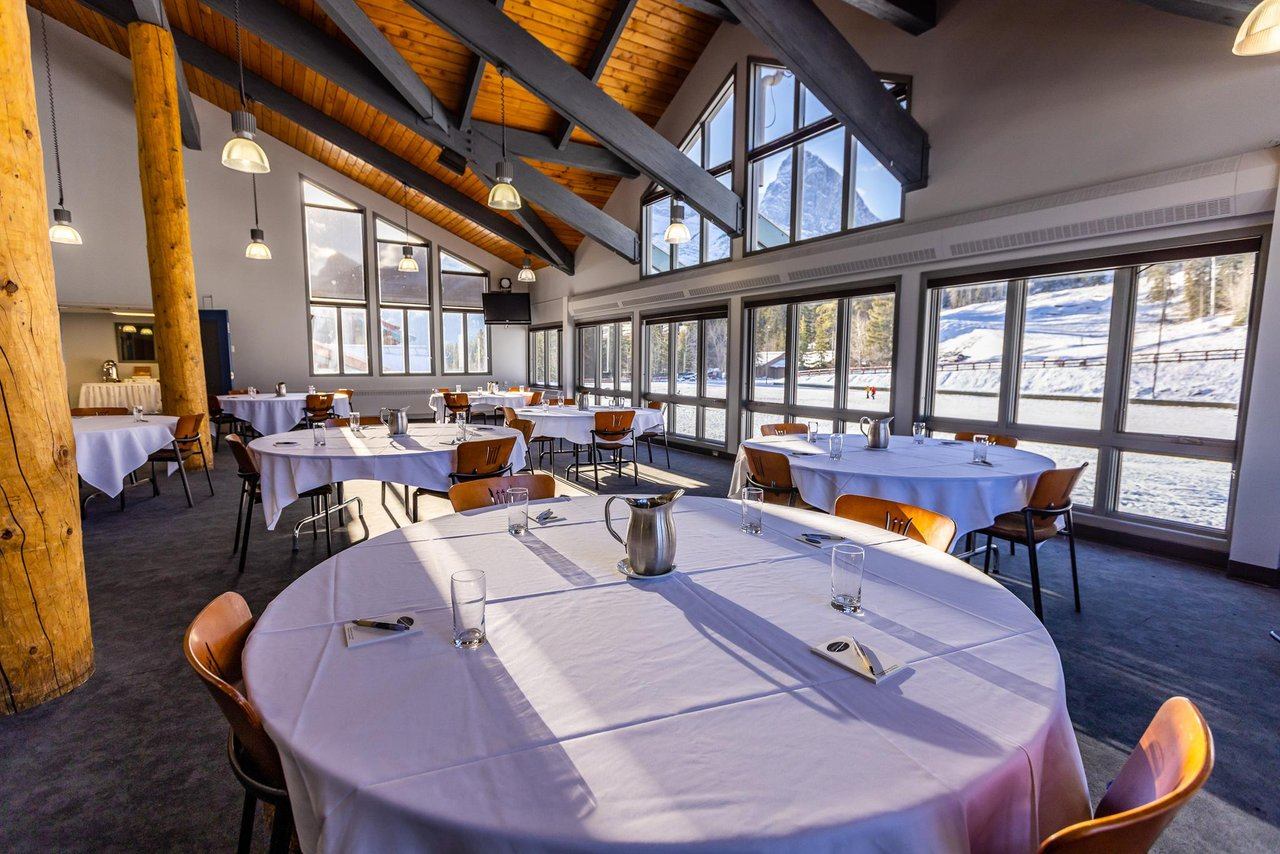 A large event room with round tables, a vaulted wooden ceiling, and expansive windows overlooking a snowy mountain landscape.