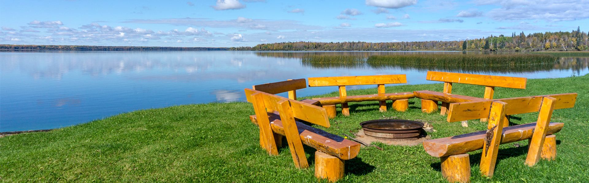Wooden benches circle a fire pit on grassy shoreline beside calm water at Iosegun Lake.