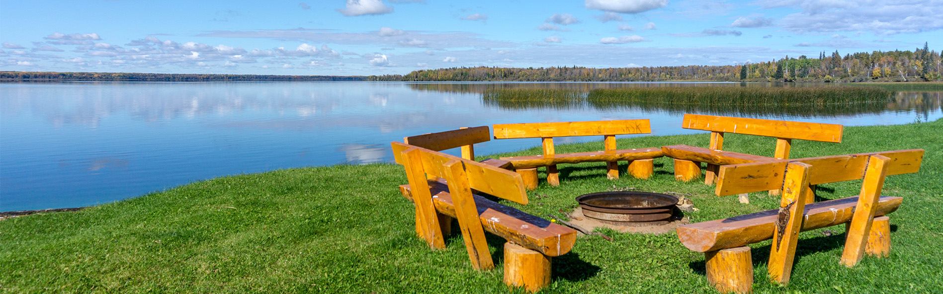 Wooden benches circle a fire pit on grassy shoreline beside calm water at Iosegun Lake.