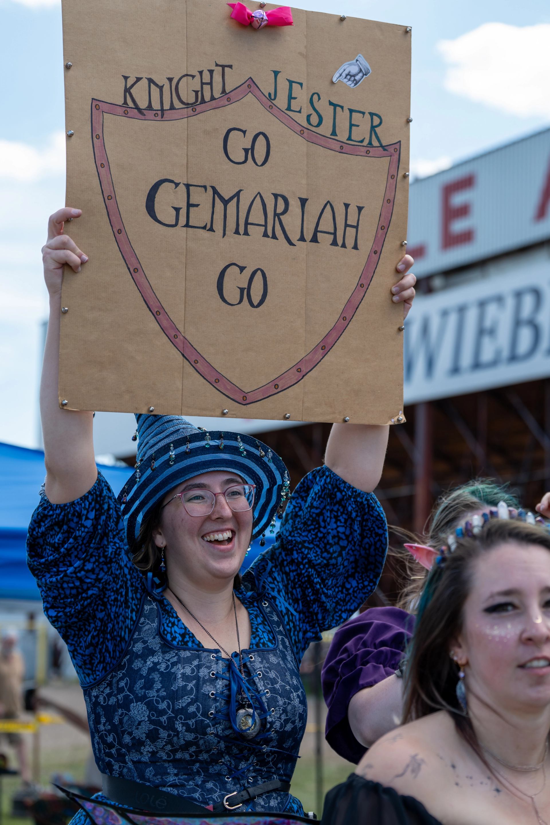 Festival attendee holding a handmade sign among a crowd at an outdoor medieval-themed event.