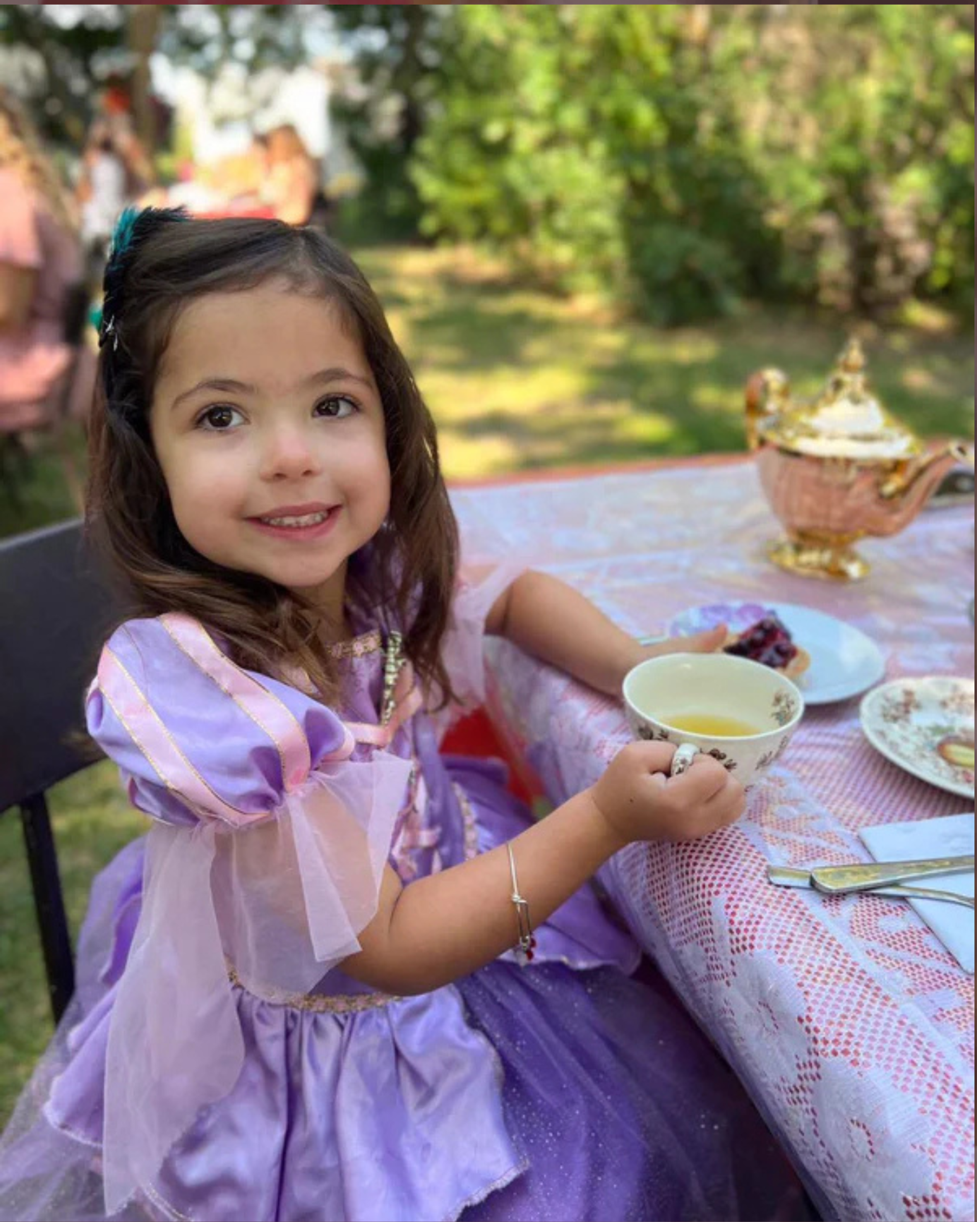Child in purple costume holding teacup at outdoor table with teapot and dessert.