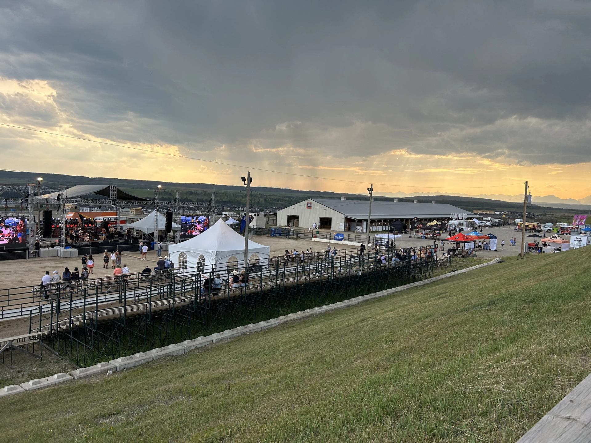 Outdoor event setup with stage, tents, and crowd near arena