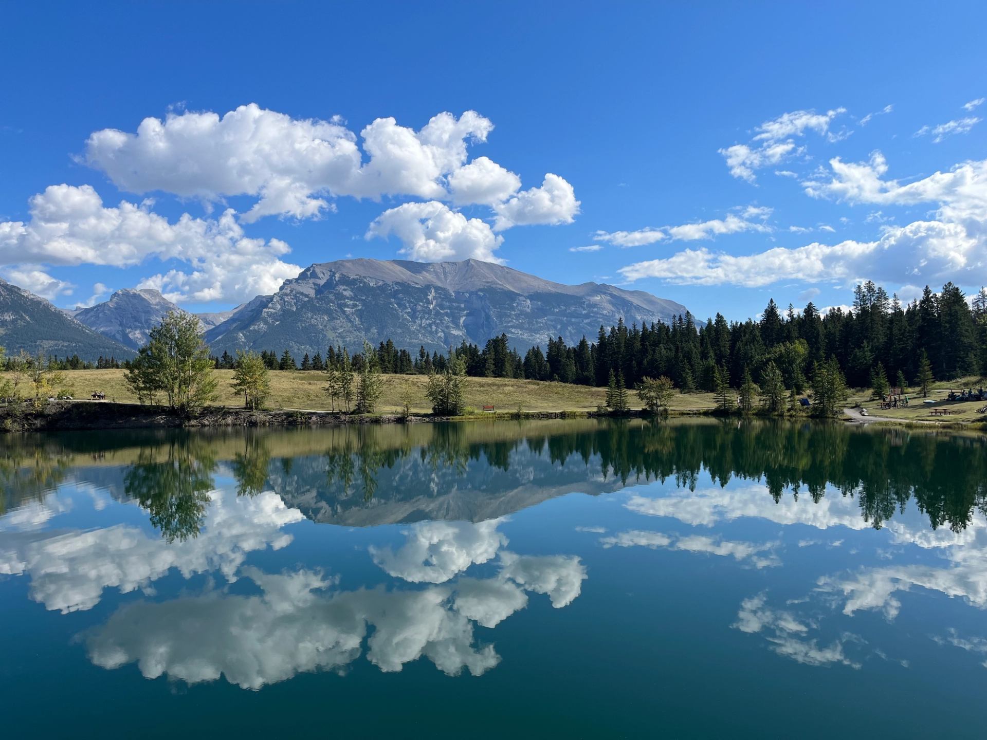 Mountain range and forest reflected in calm lake under blue sky with scattered clouds.