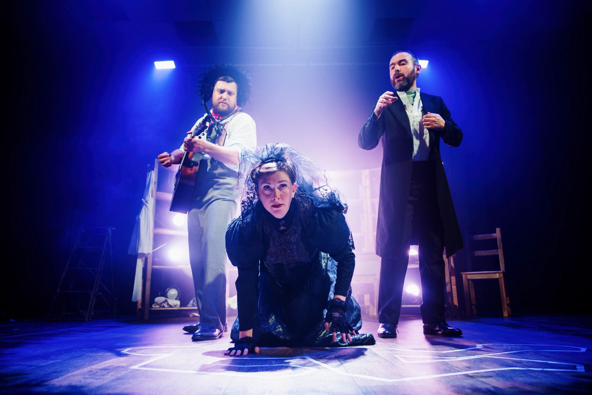 Stage performance with costumed actors, one crouching front, dramatic blue and purple lighting.