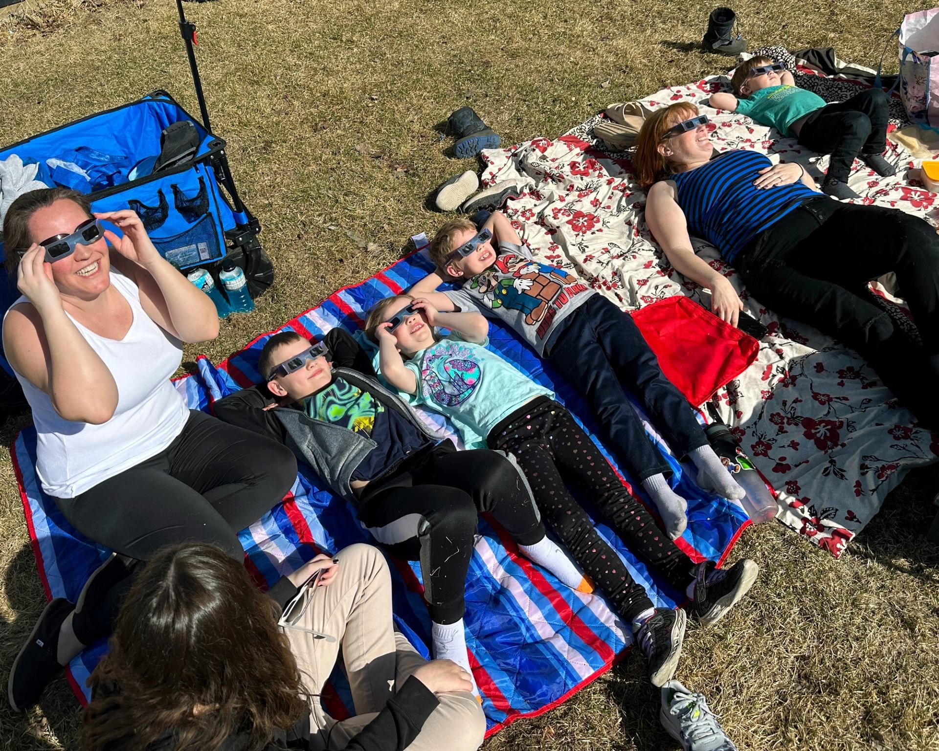 Group lying on blankets outdoors wearing eclipse glasses and looking up at the sky.