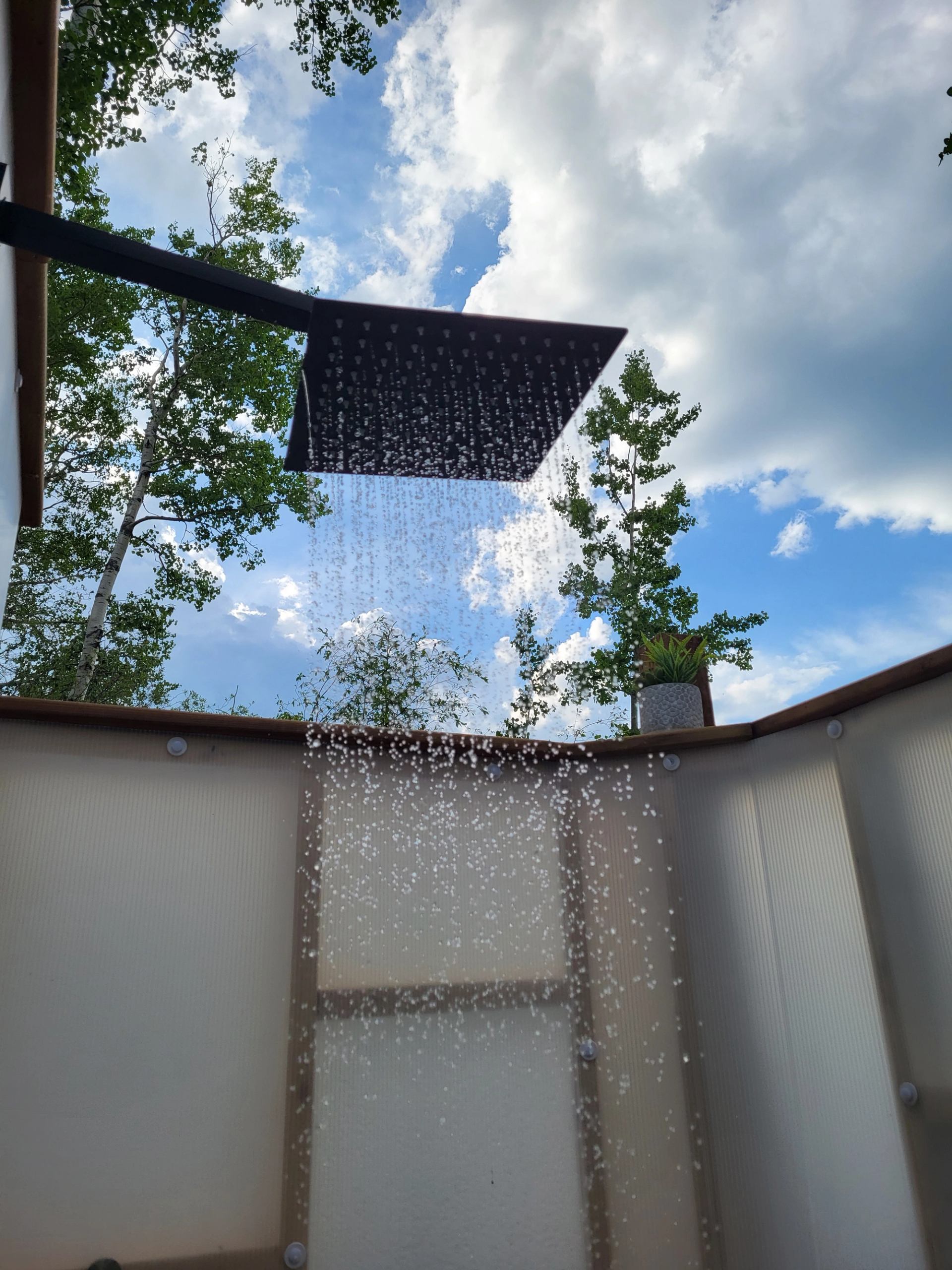 An outdoor shower with water spraying under a blue, cloudy sky and trees.