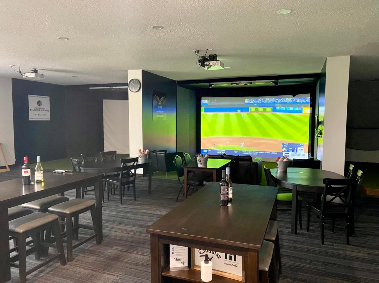 Indoor golf simulator lounge with tables, chairs, and a screen showing a course.