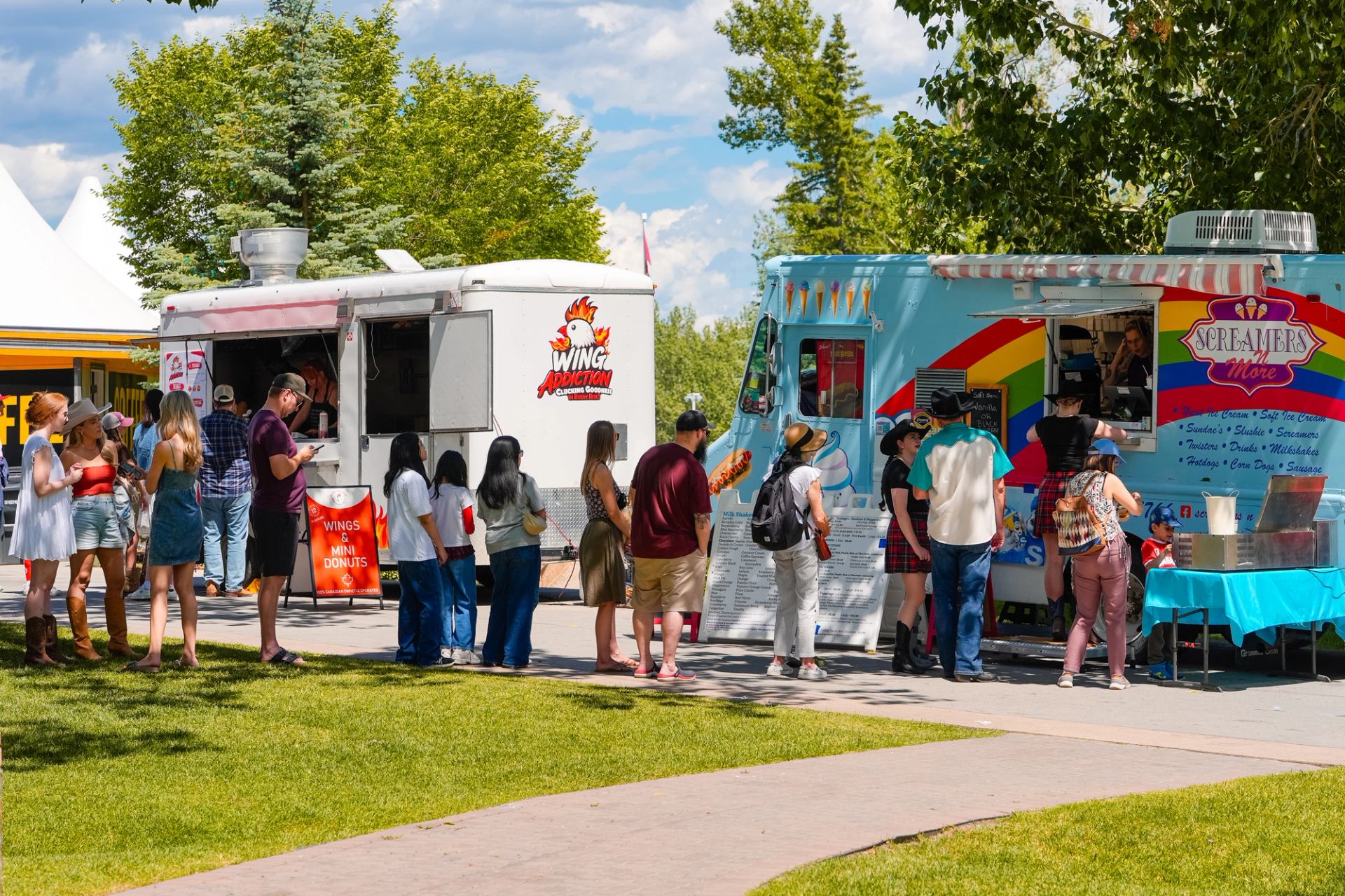 People line up at colourful food trucks in a park with green trees.