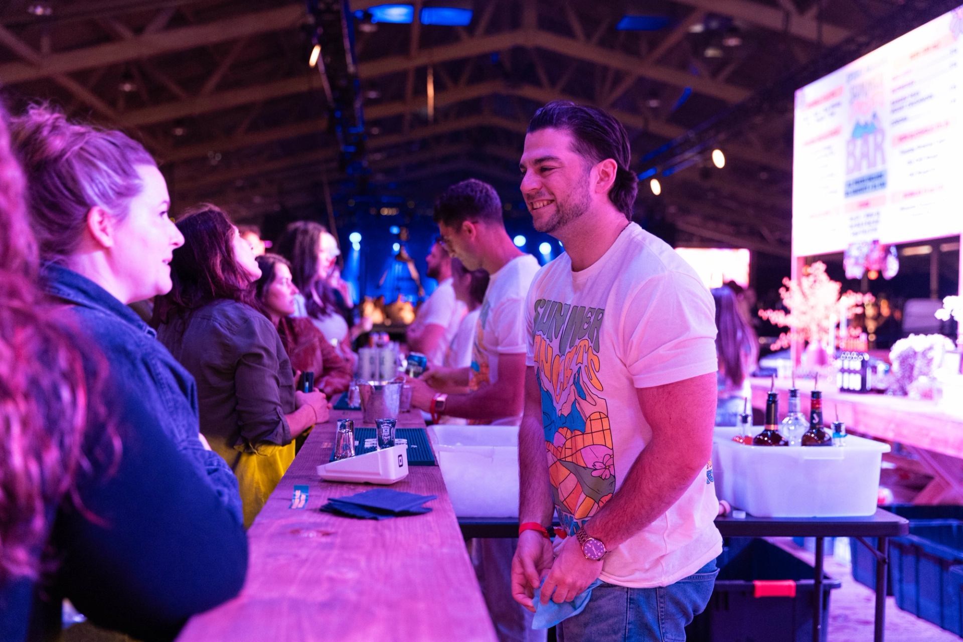 Crowded bar counter with people ordering drinks from staff under purple event lighting.