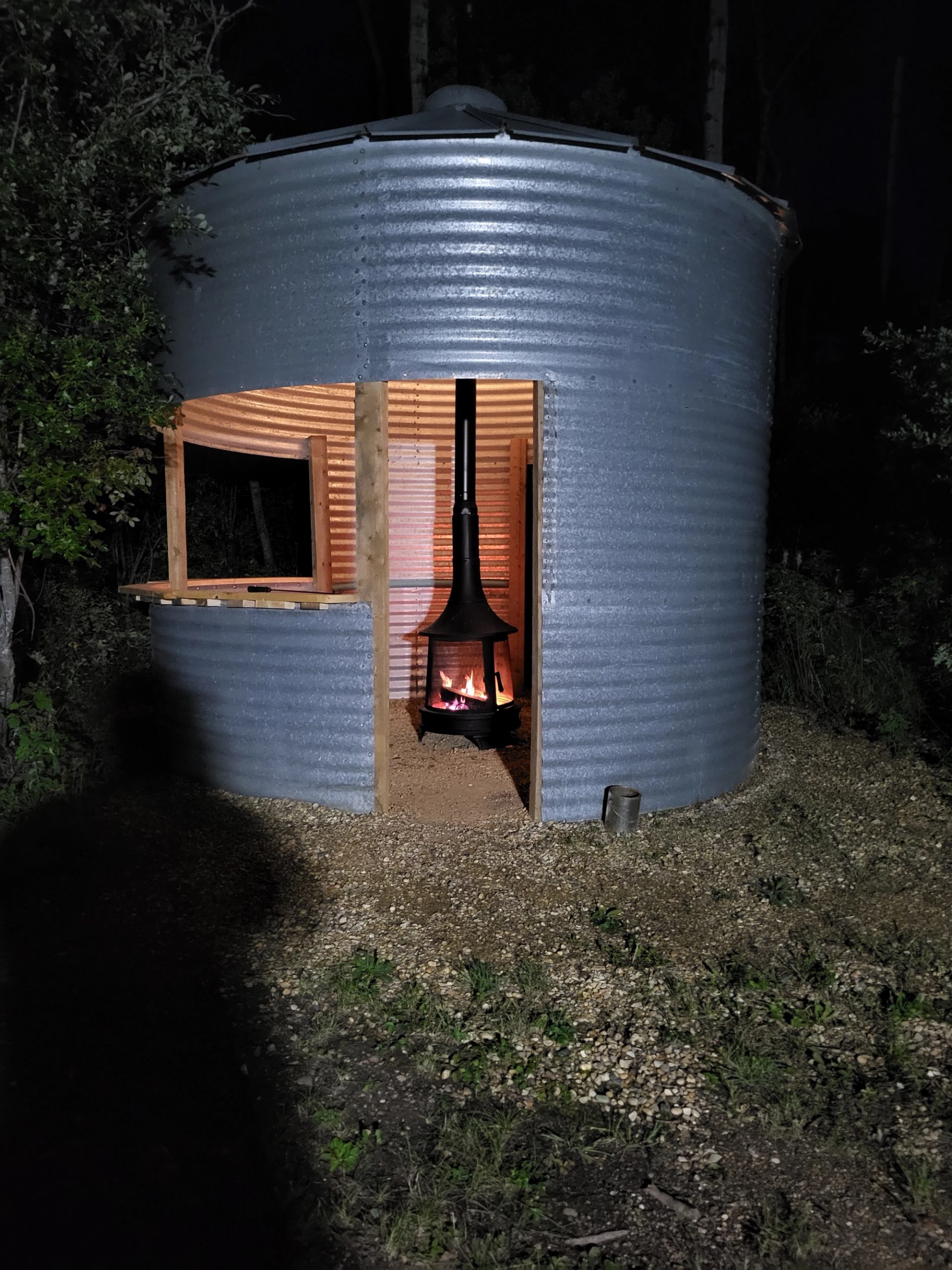 A repurposed corrugated metal grain silo with a fire pit glowing inside at night.