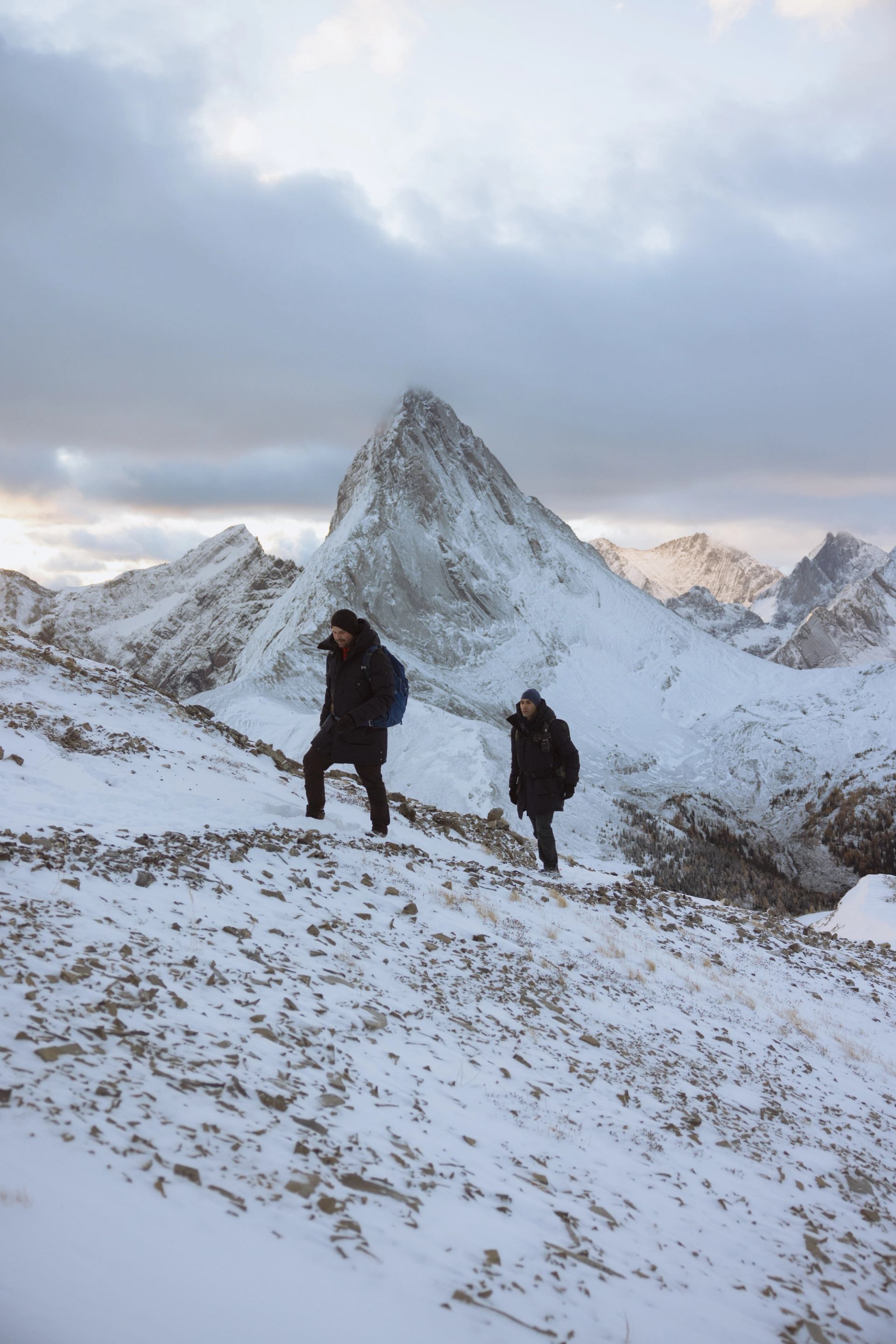 Two hikers walking along a snowy mountain ridge with dramatic peaks in the background.