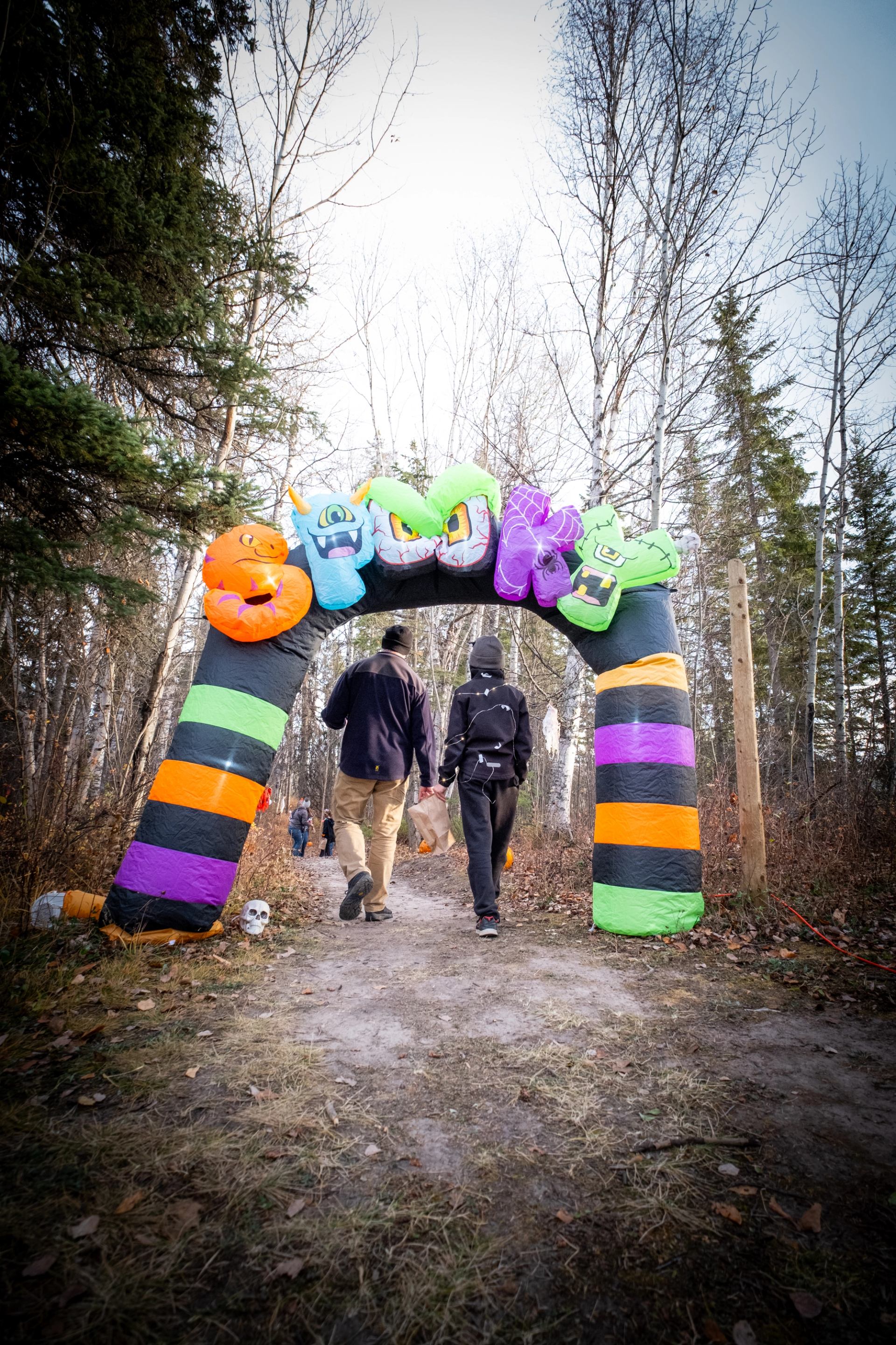 People walk under inflatable monster arch on forest trail with scattered decorations.