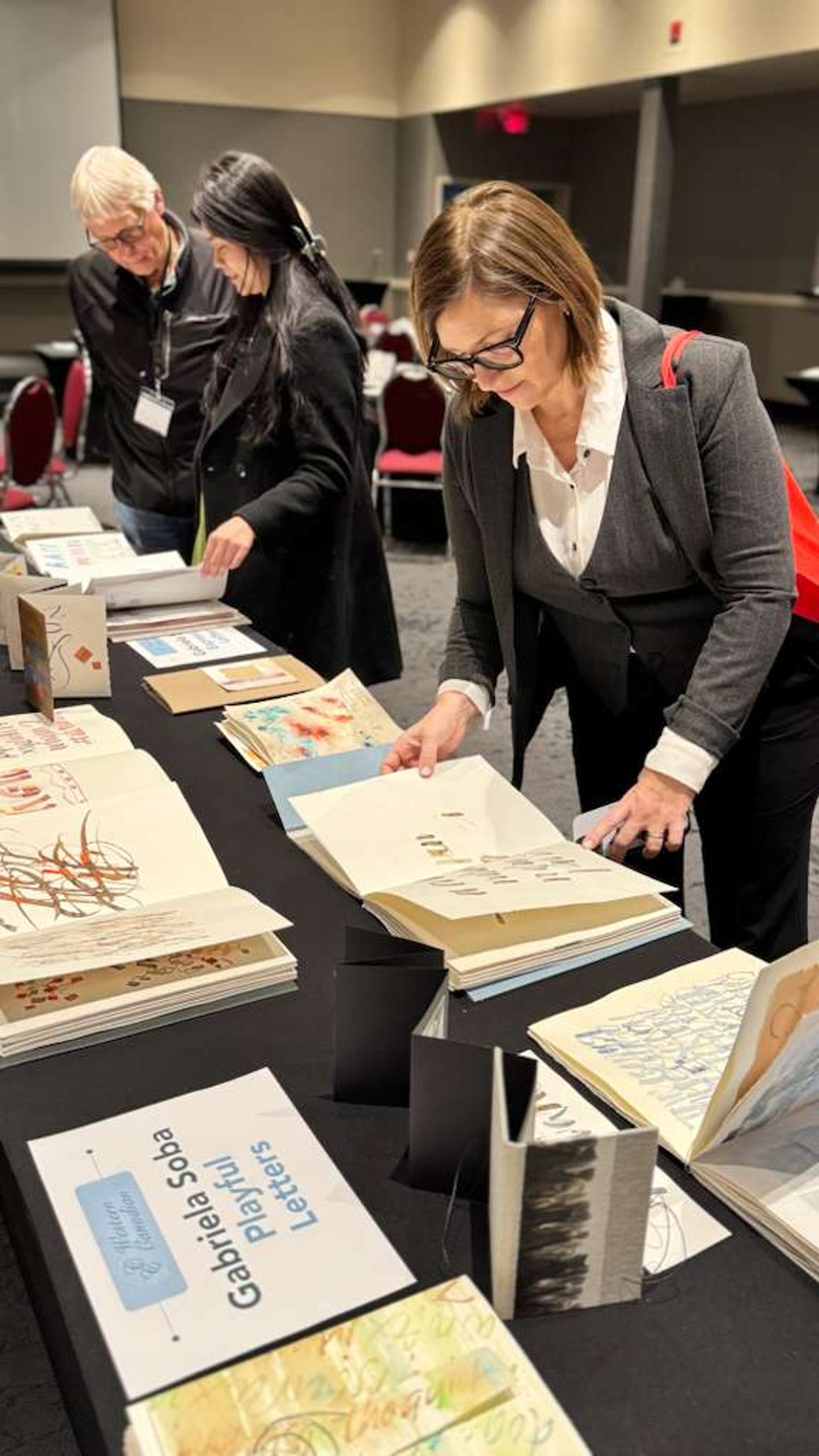 Visitors examine illustrated books and prints displayed on a table.