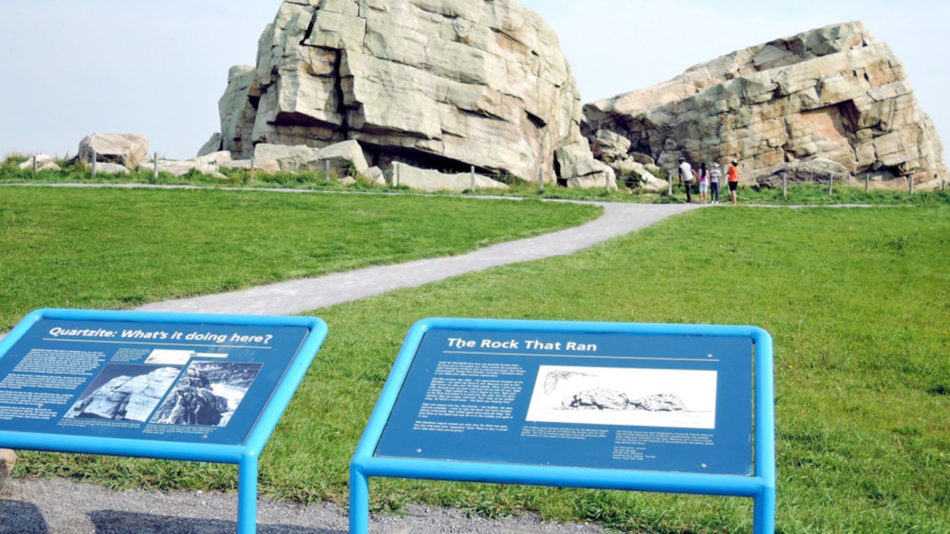 Outdoor interpretive signs about quartzite and The Rock That Ran facing large rock formations.