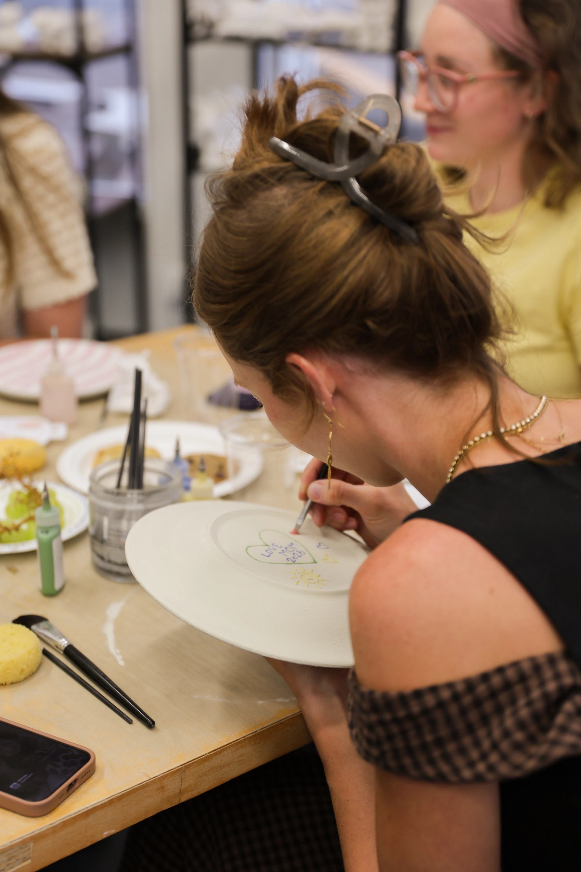 Person painting a white ceramic plate at a shared art table with supplies.