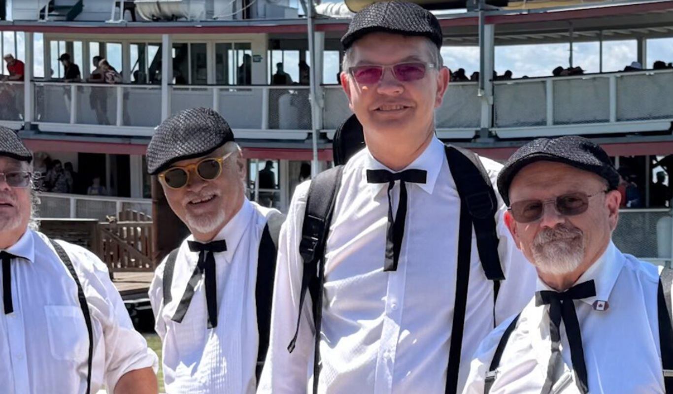 Four people in white shirts, suspenders and caps stand in front of a boat.