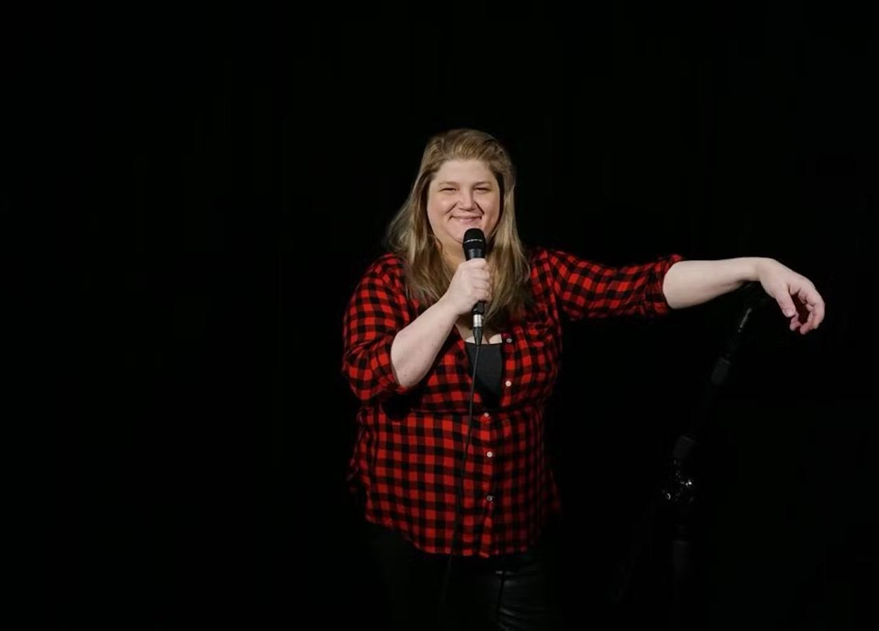 Female comedian in red-and-black plaid shirt holding microphone on dark stage backdrop.