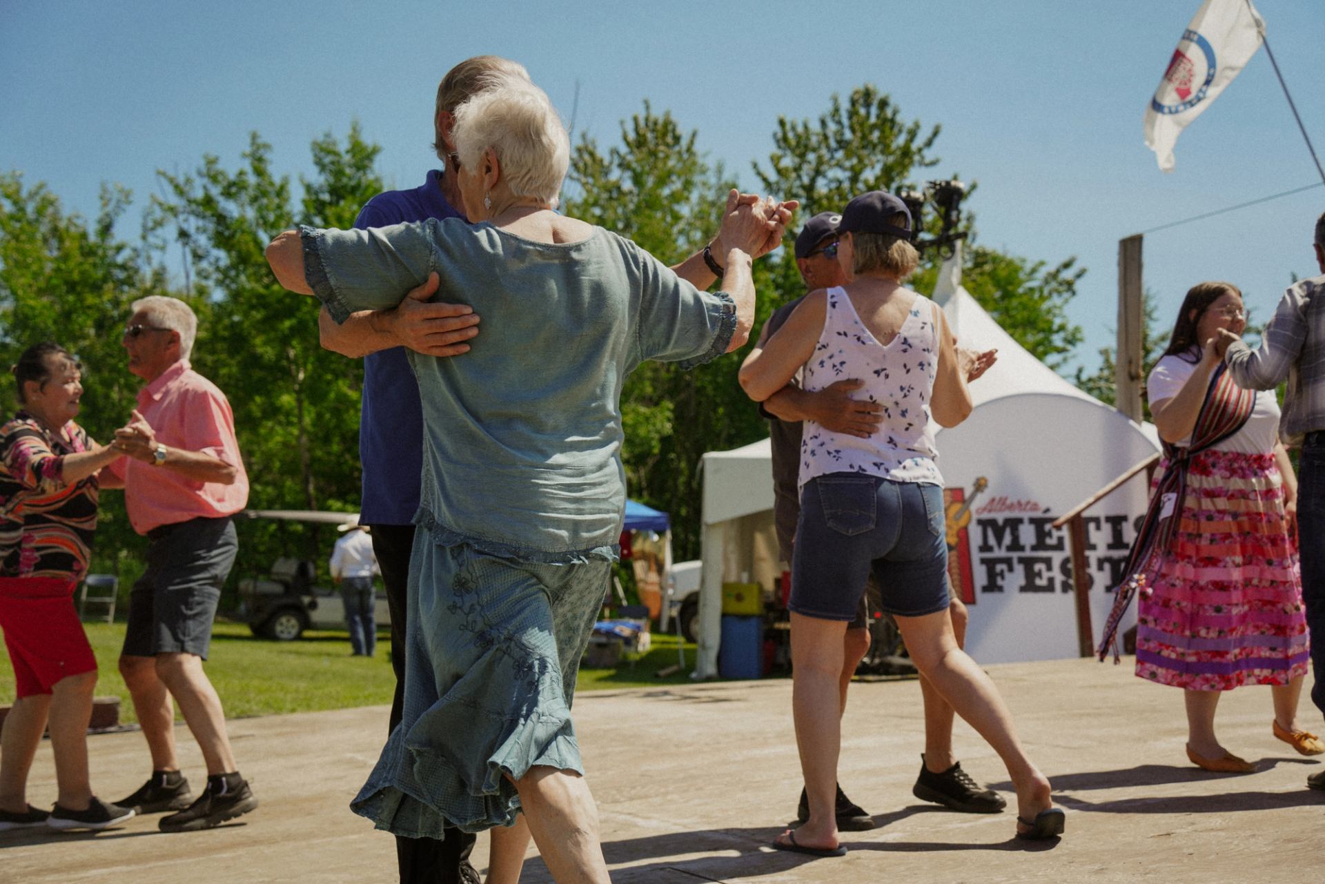 People dance together on an outdoor wooden stage at a Métis festival.
