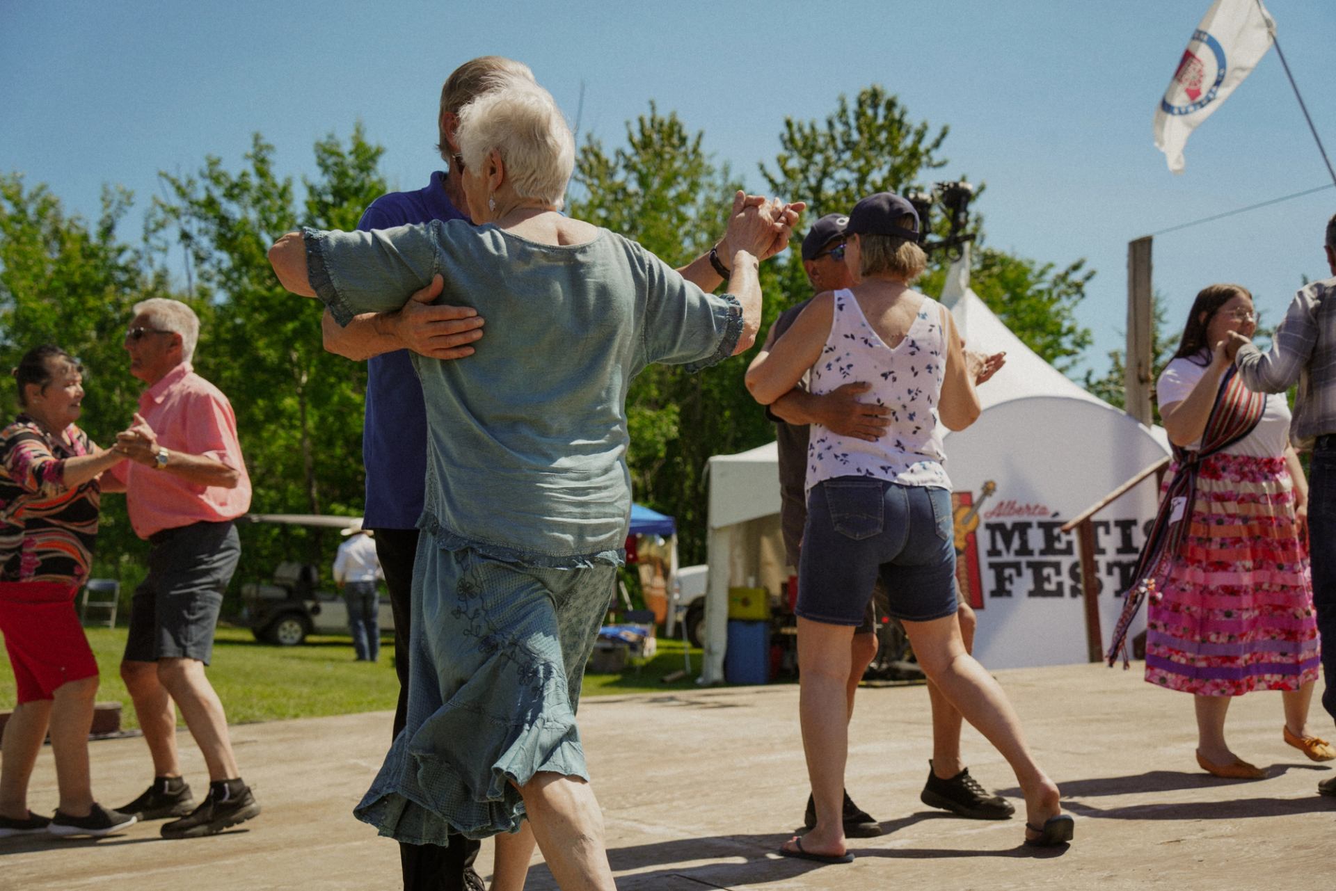 People dance together on an outdoor wooden stage at a Métis festival.