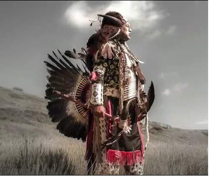 Person in feathered regalia stands in prairie grass with hills and cloudy sky.