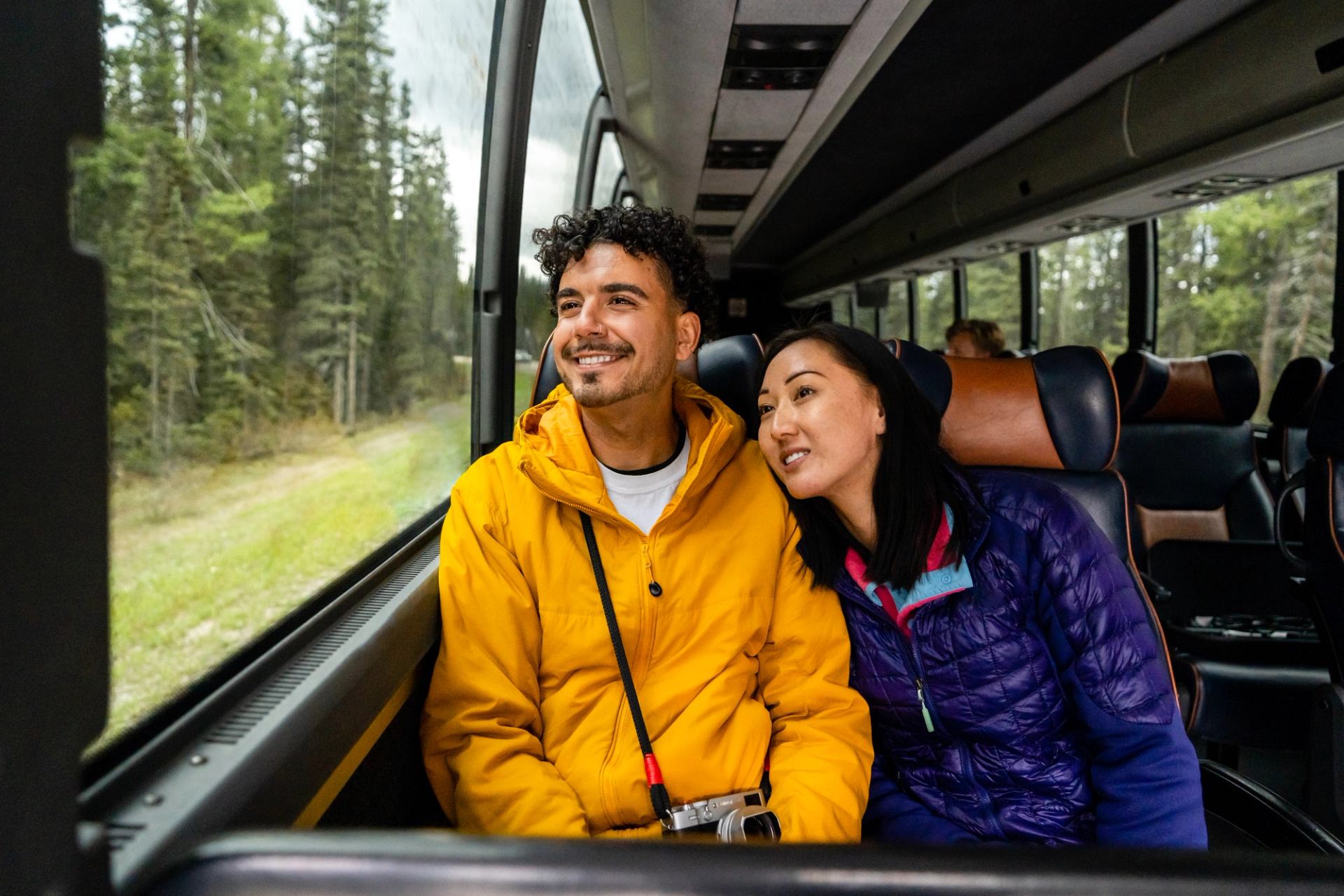 Two passengers seated together on a shuttle bus, wearing colorful outdoor jackets, looking out the window at a dense green forest; one passenger has a camera resting on their lap, and the bus interior features large windows and cushioned seats.